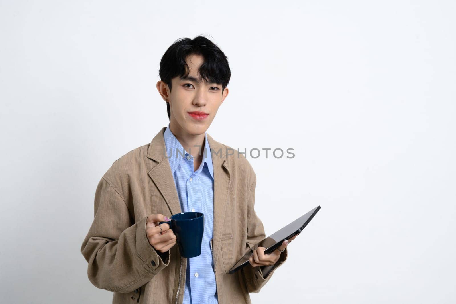 Confident young businessman holding a coffee mug and digital tablet standing over white background.