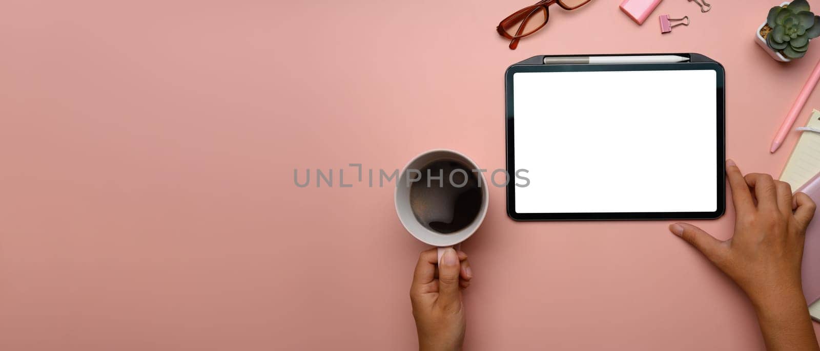 Overhead view of hand holding cup of coffee and using digital tablet on pink pastel background with copy space.