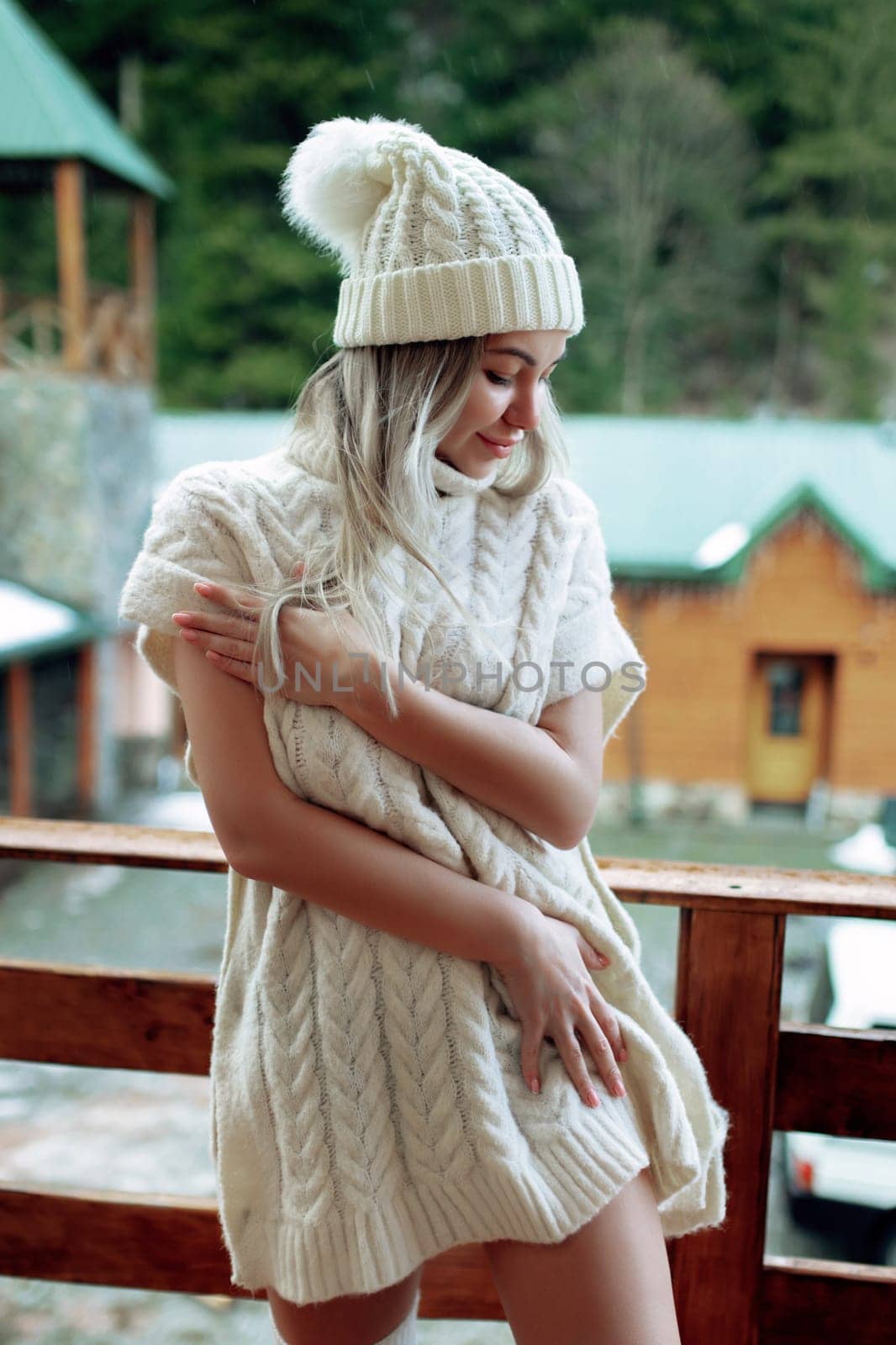 A sexy girl in a white sweater and stockings on the balcony admires the beauty of nature. wool clothes and a hat on a beautiful blonde.