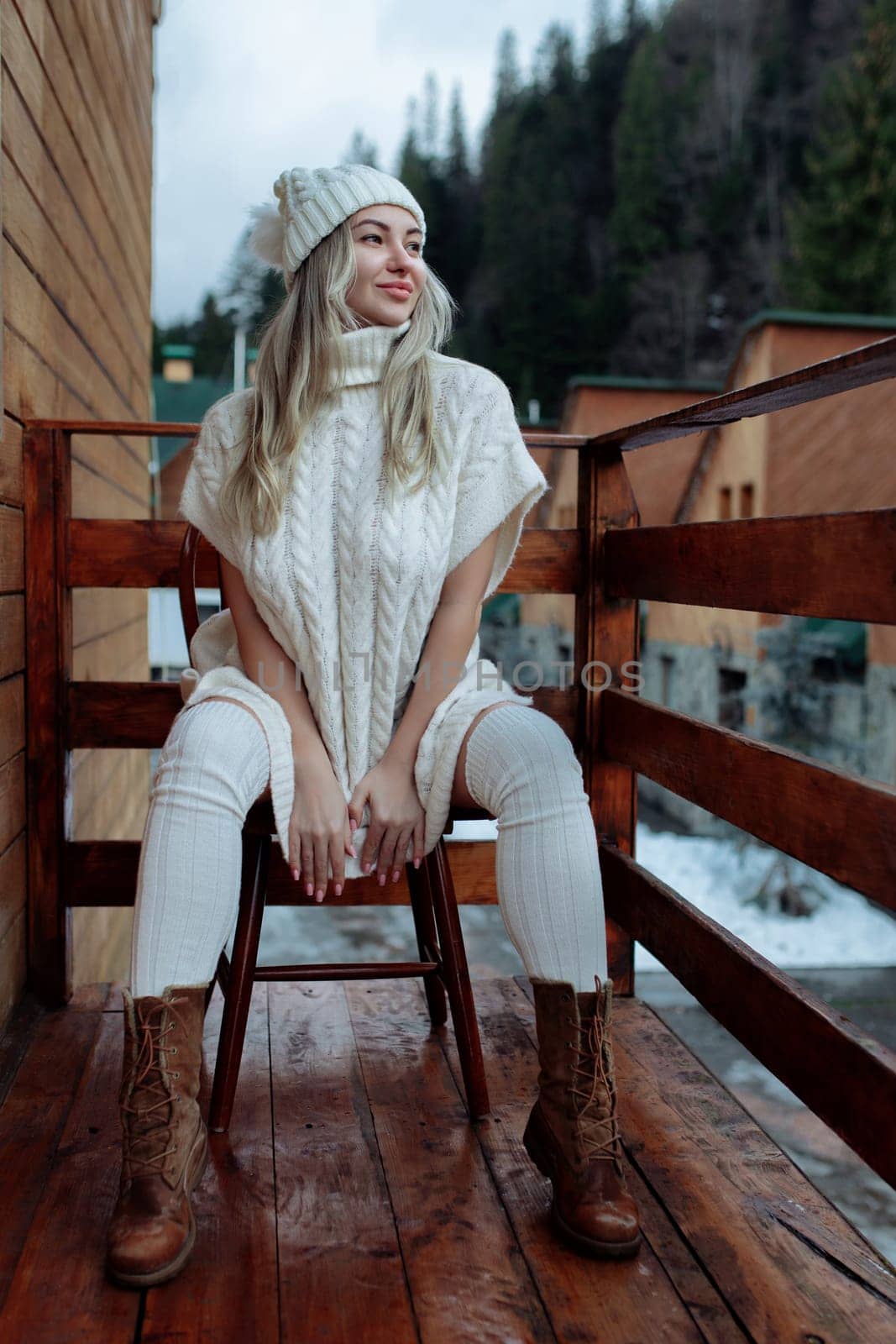 A sexy girl in a white sweater and stockings on the balcony admires the beauty of nature. wool clothes and a hat on a beautiful blonde.