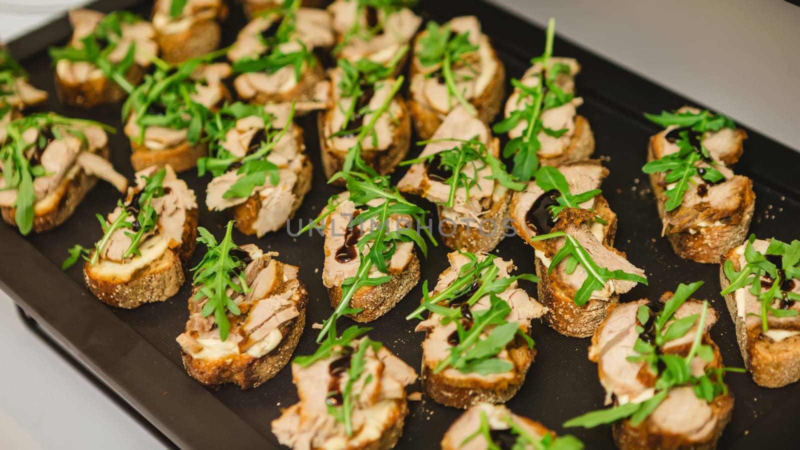 A tray of gourmet bruschetta appetizers featuring slices of bread topped with meat, arugula, and a drizzle of balsamic glaze, perfect for parties or gatherings.
