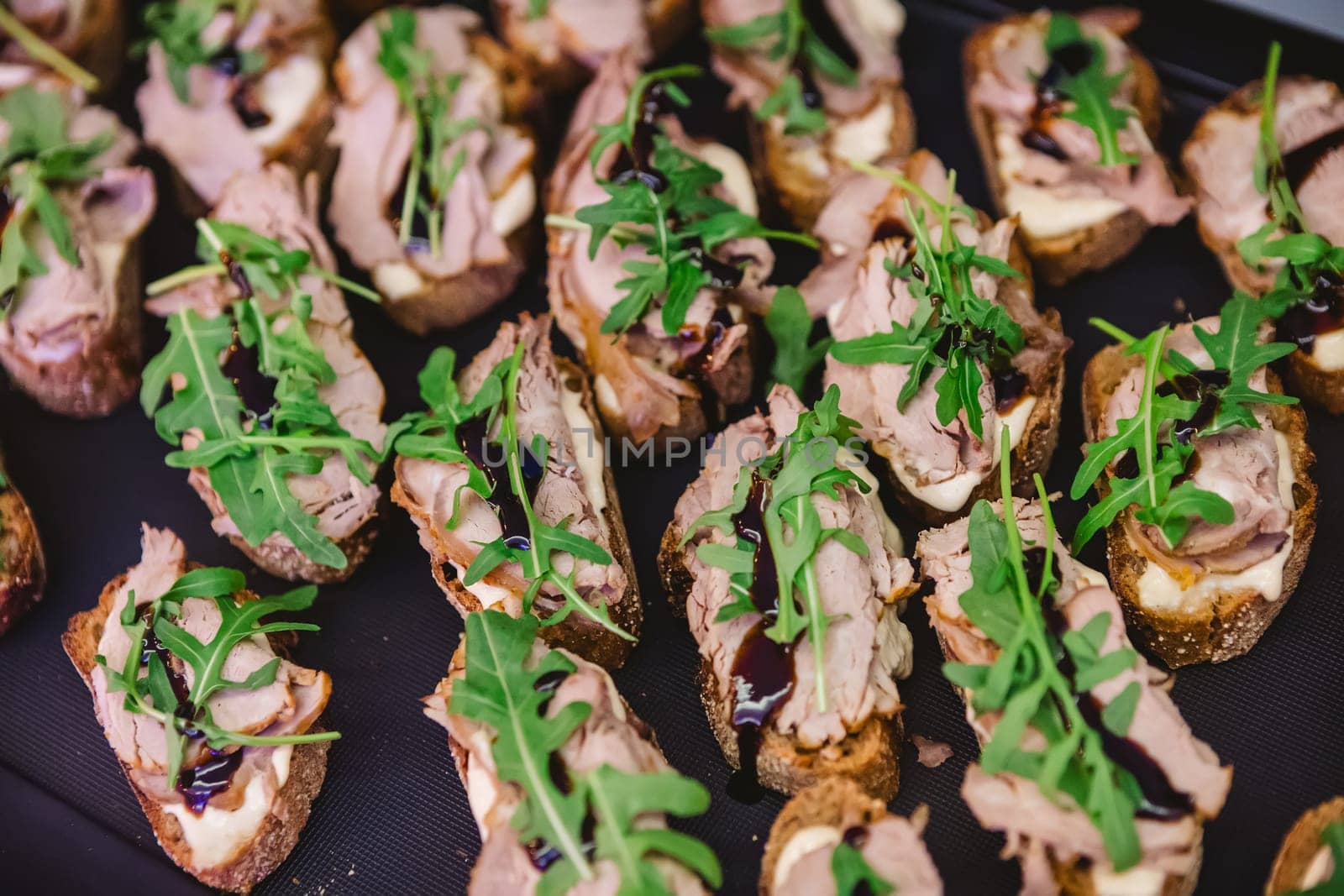A tray of gourmet bruschetta appetizers featuring slices of bread topped with meat, arugula, and a drizzle of balsamic glaze, perfect for parties or gatherings.