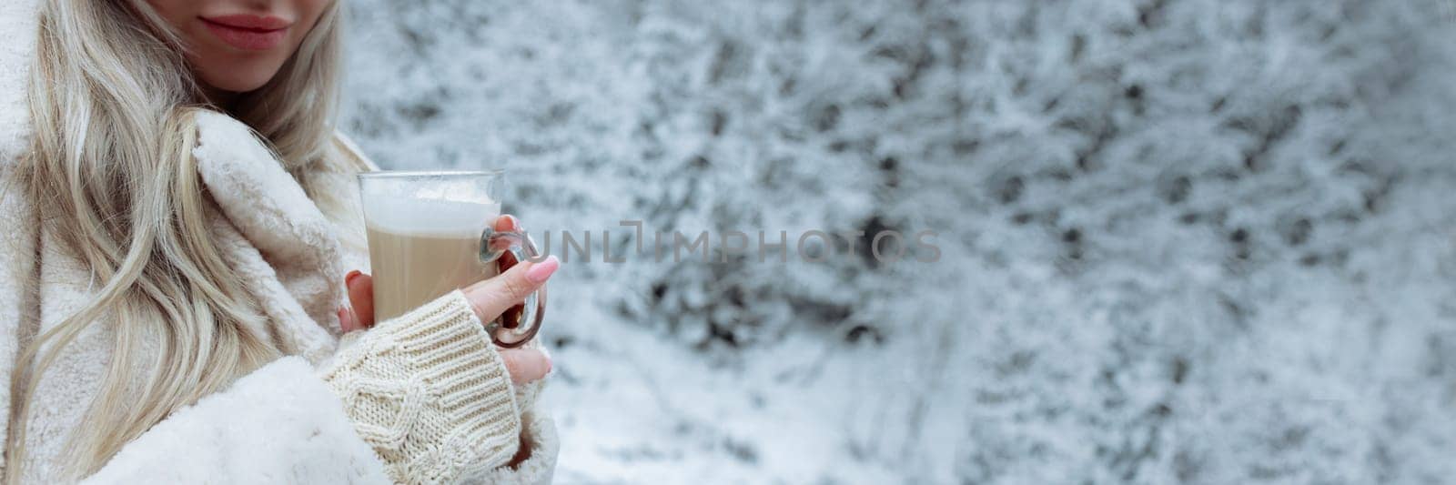 A beautiful blonde in winter clothes holds a cappuccino mug in her hands. A place for text gentle snow background.