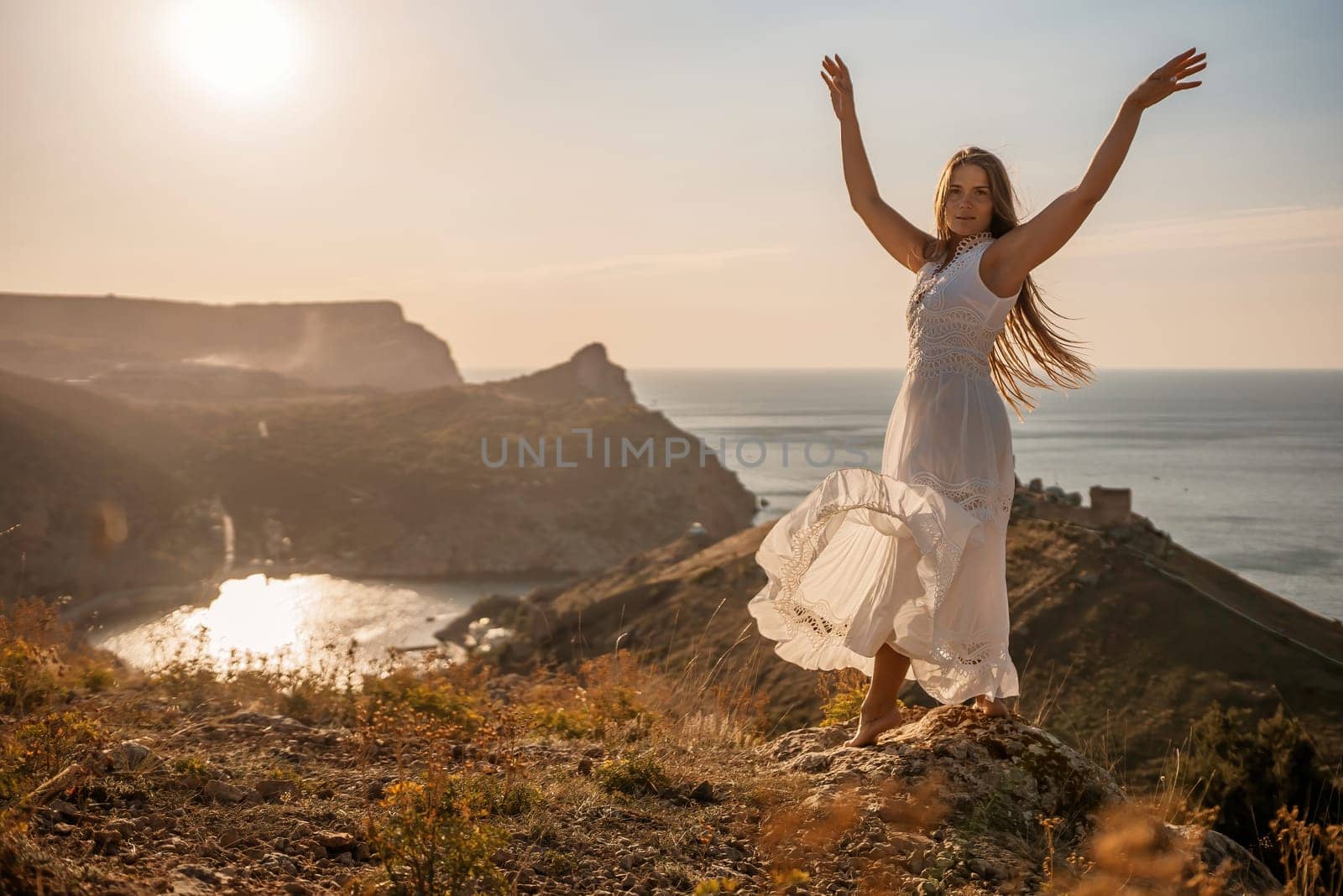 A woman stands on a hill overlooking the ocean. She is wearing a white dress and has long hair. The scene is serene and peaceful, with the sun shining brightly in the background