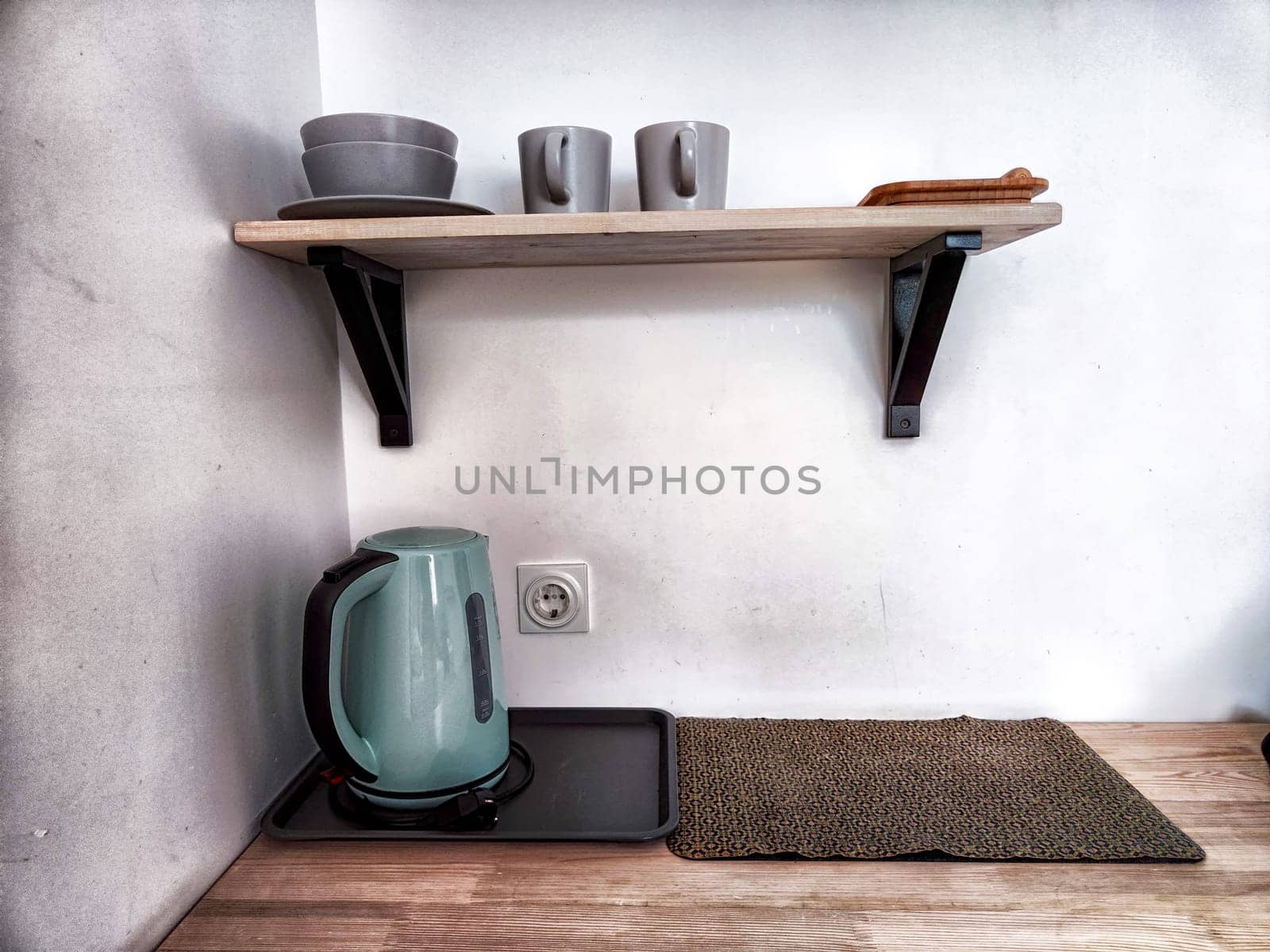 A green electric kettle sits on wooden countertop below a shelf holding two gray mugs and a small wooden tray