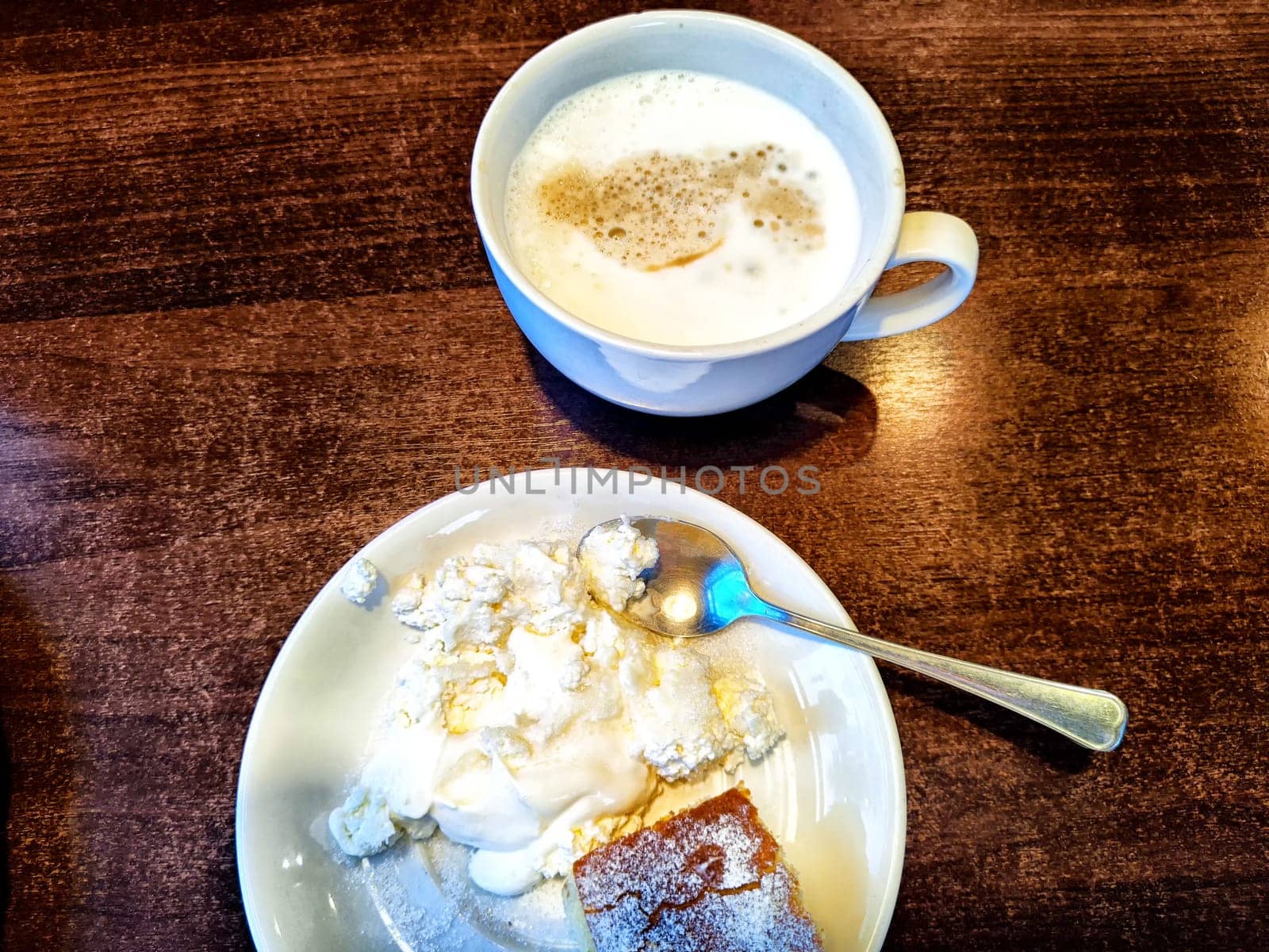 A cup of frothy coffee sits beside a plate of creamy dessert and cake, all arranged on a rustic wooden table.