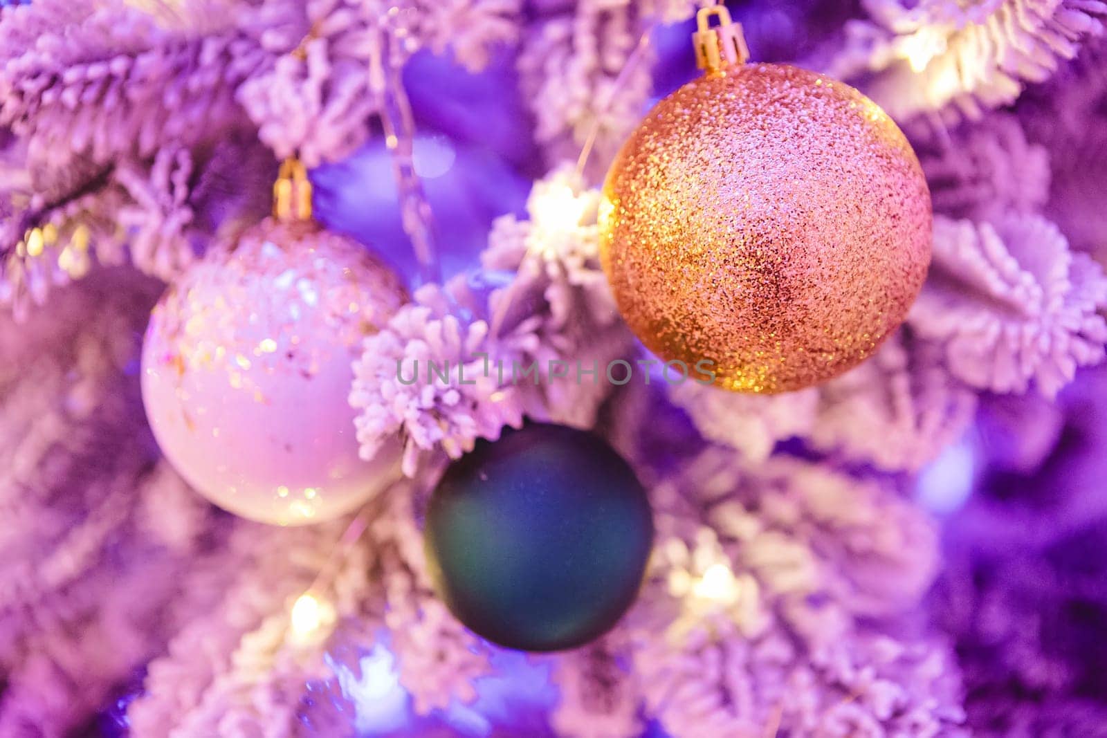 A close-up of a Christmas tree adorned with glittery ornaments. The image features a gold glitter ball, a white ornament with gold specks, and a dark green bauble, all hanging on frosted branches.