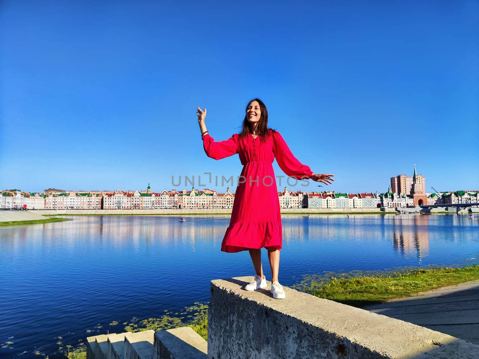 A woman smiles joyfully while standing on a concrete ledge near a river, showcasing vibrant architecture and clear blue sky.