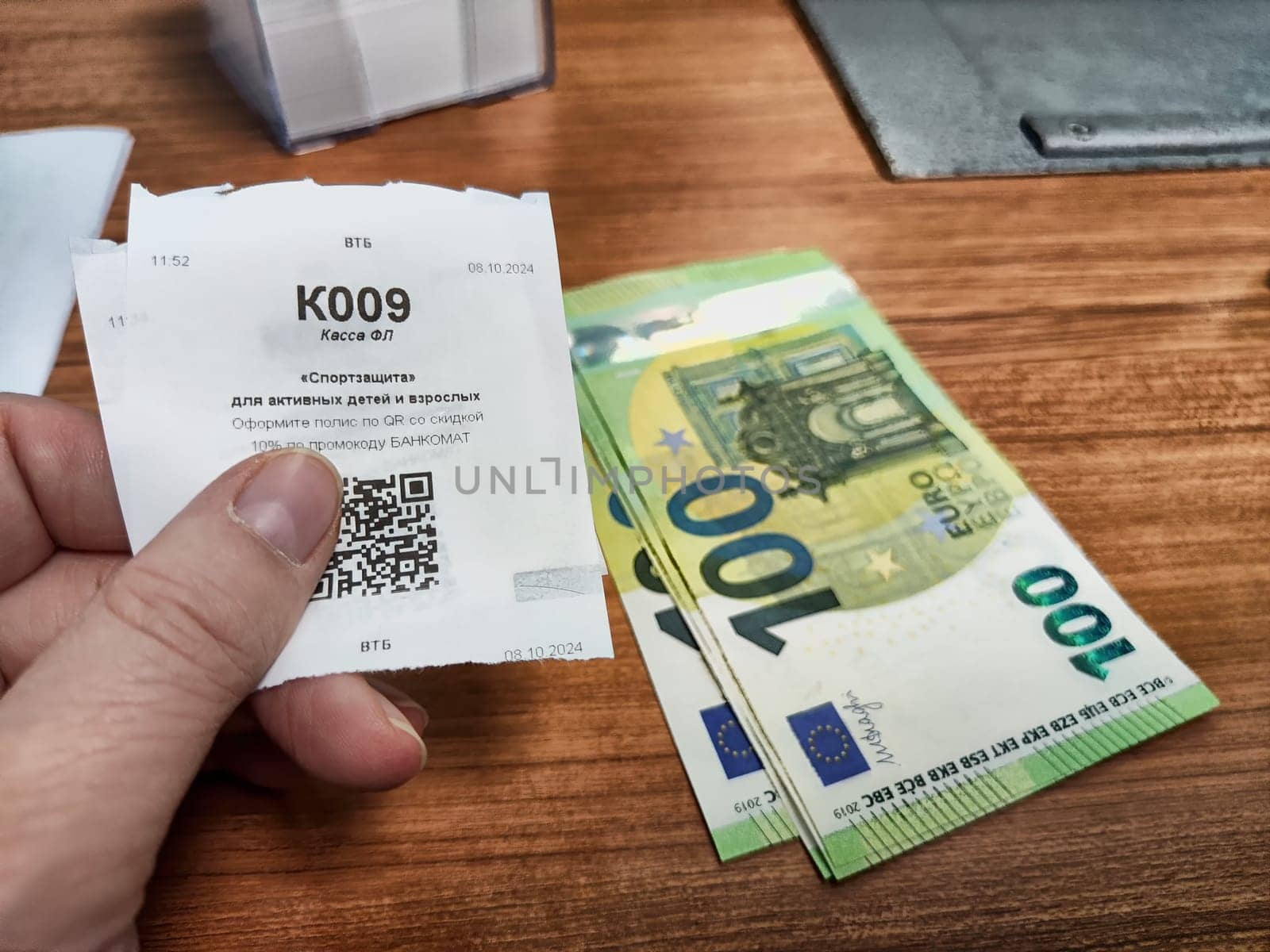 Kirov, Russia - October 08, 2024: A person holding a voucher for currency exchange next to a stack of 100 euro banknotes at a bustling exchange counter.