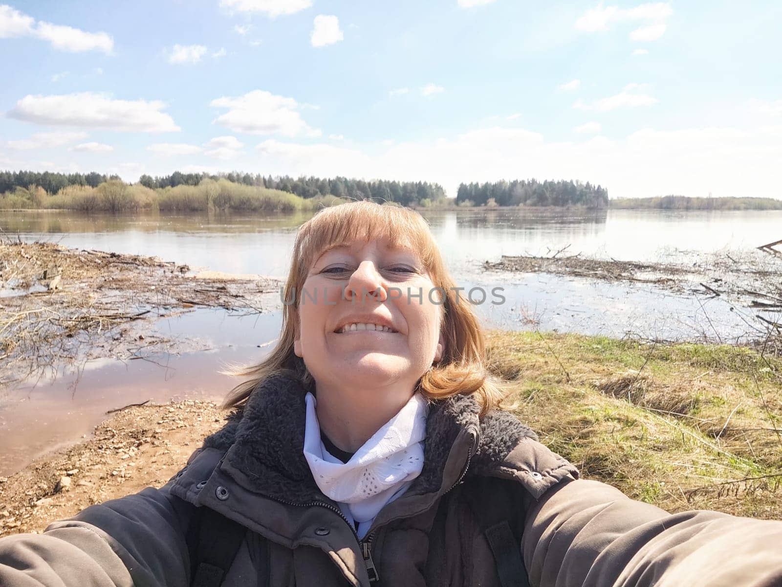 A woman in her 40s or 50s takes selfie outdoors near water on a sunny day. She is wearing a blue jacket and smiling brightly at the camera