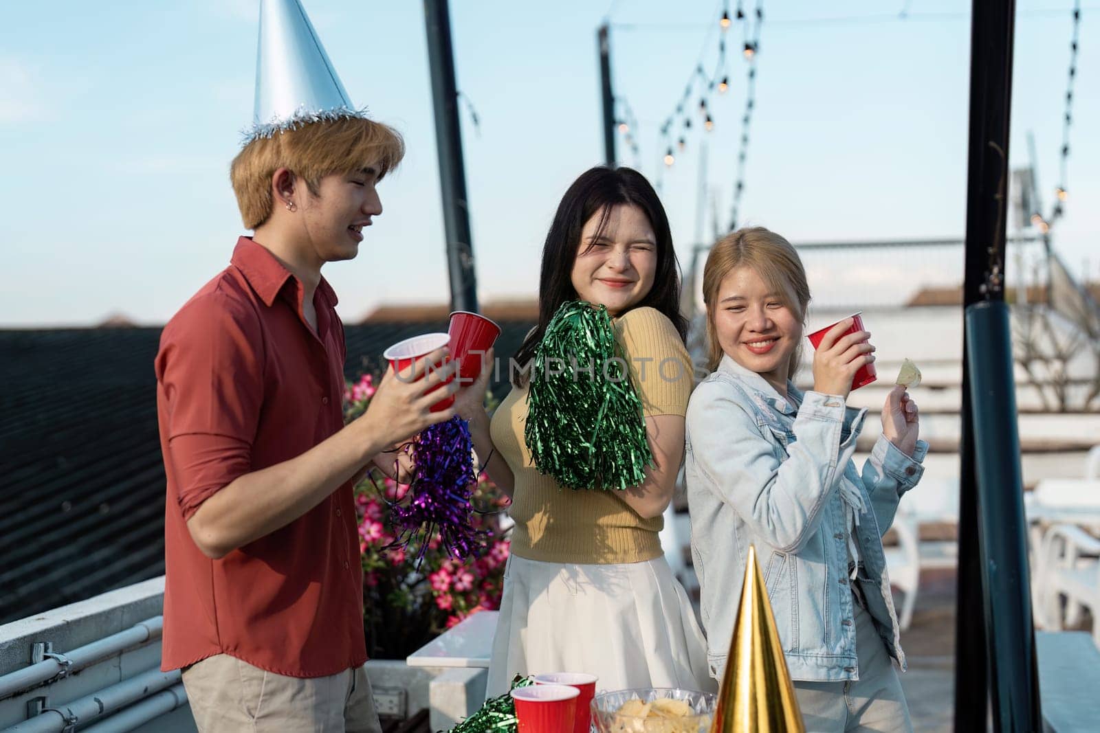 Friends enjoying a lively rooftop party with drinks, festive hats, and string lights on a sunny day.