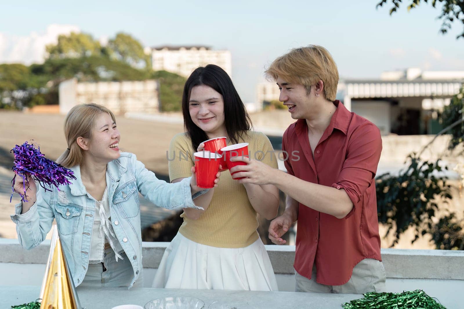 Three friends enjoying a lively rooftop party, clinking red cups and celebrating with colorful decorations under a clear sky.