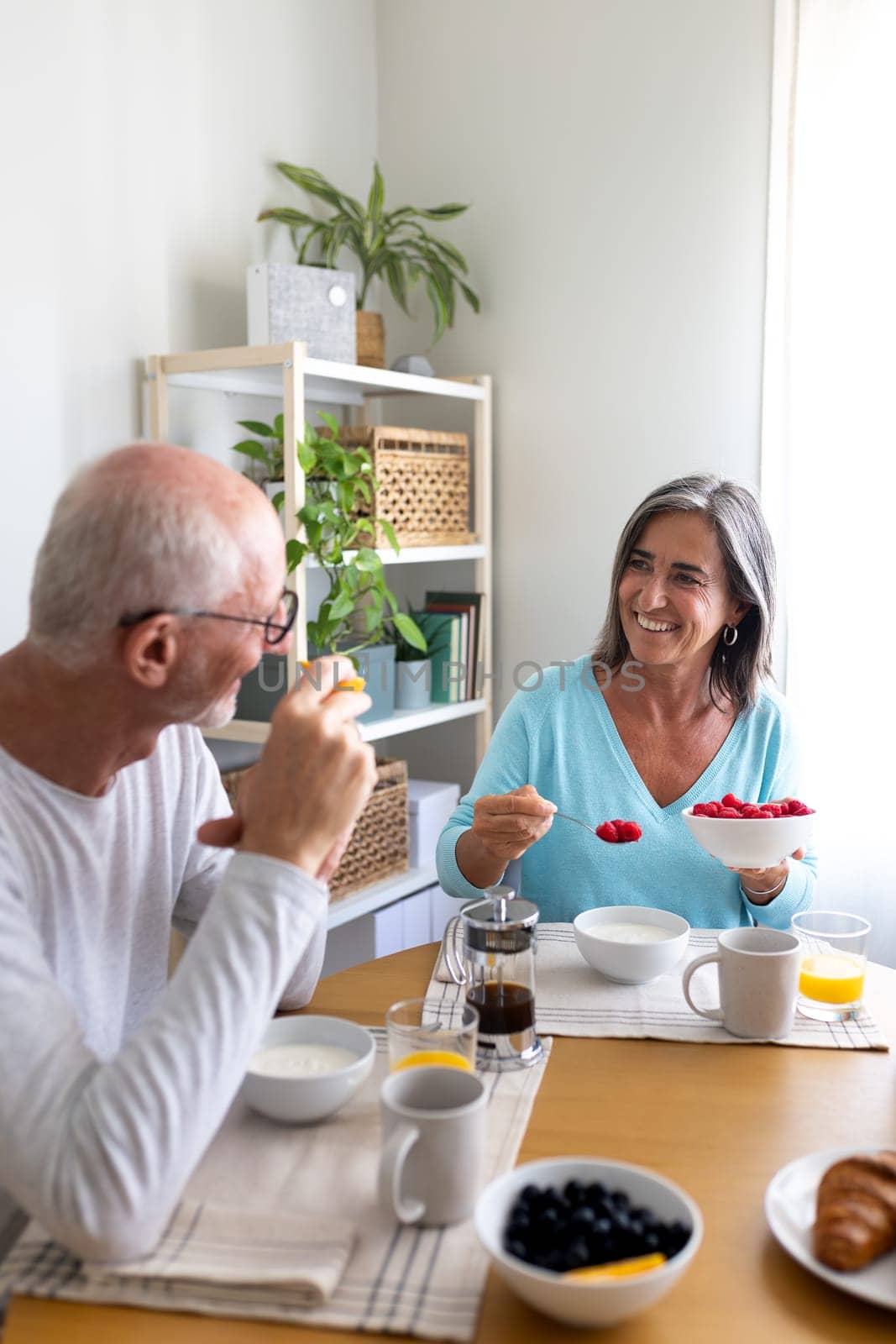 Vertical portrait of happy mature couple having healthy breakfast at home. Man eating orange, woman serving raspberries. Lifestyle concept.