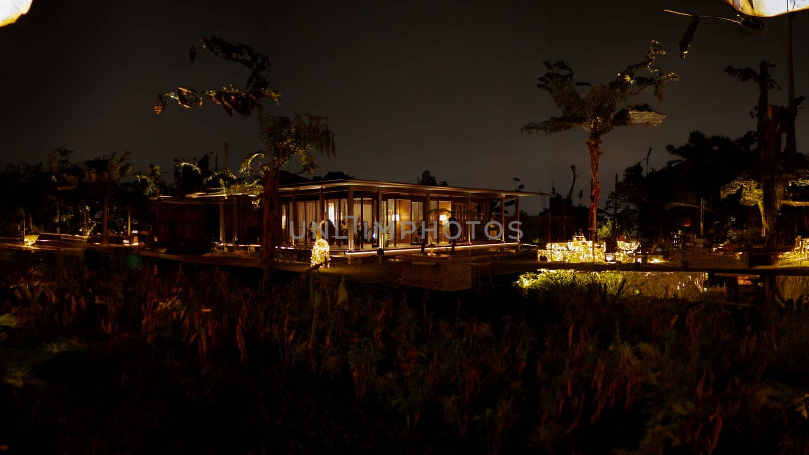 Tropical resort villa at night with illuminated interior, surrounded by lush vegetation under a starry sky.