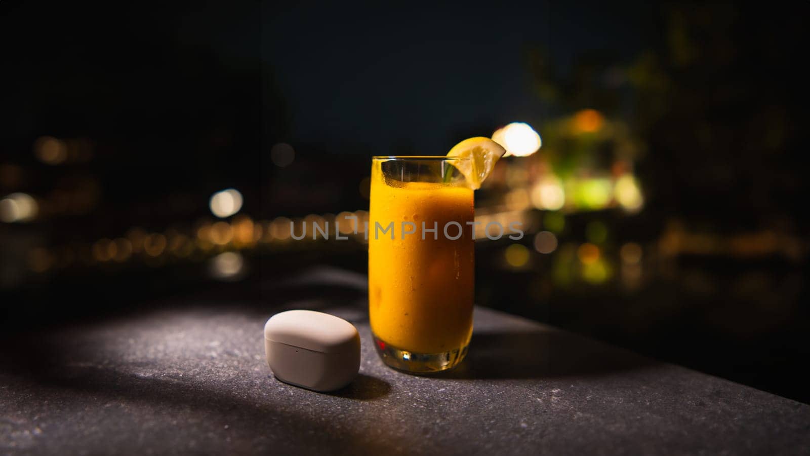 Glass of orange juice with a slice of lemon on a table at night, with blurred lights in the background.