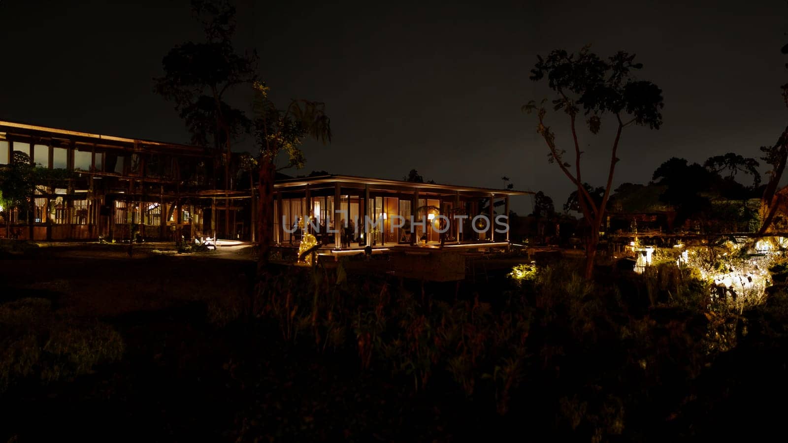 Nighttime view of a modern house with illuminated interiors and exterior lights, surrounded by dark silhouettes of trees in the dark night