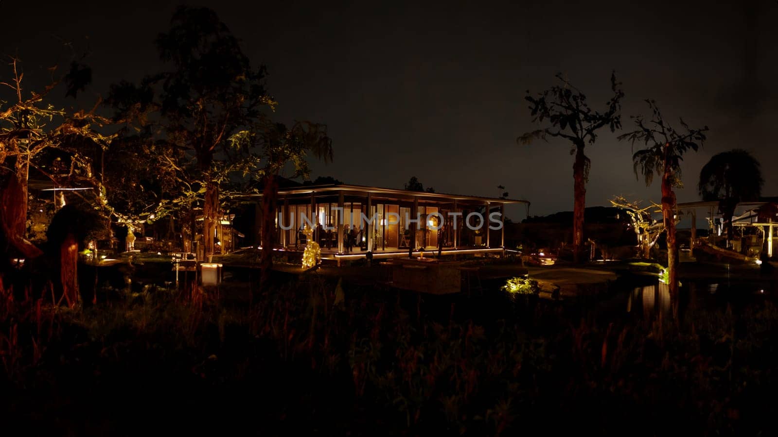 Luxurious house illuminated at night with warm lights, surrounded by trees and a calm water body, reflecting a serene ambiance in the dark night