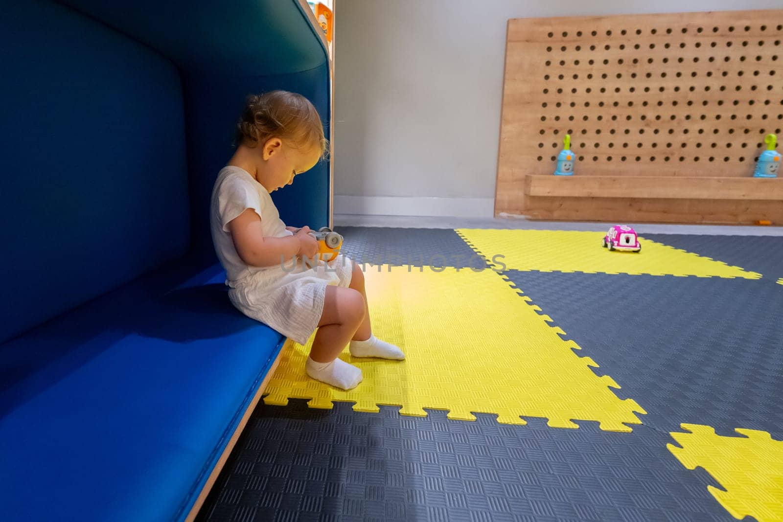 Small child sits on a cozy bench in a colorful playroom, holding a toy and exploring the environment. Concept of joyful play, early education, and creative spaces for kids