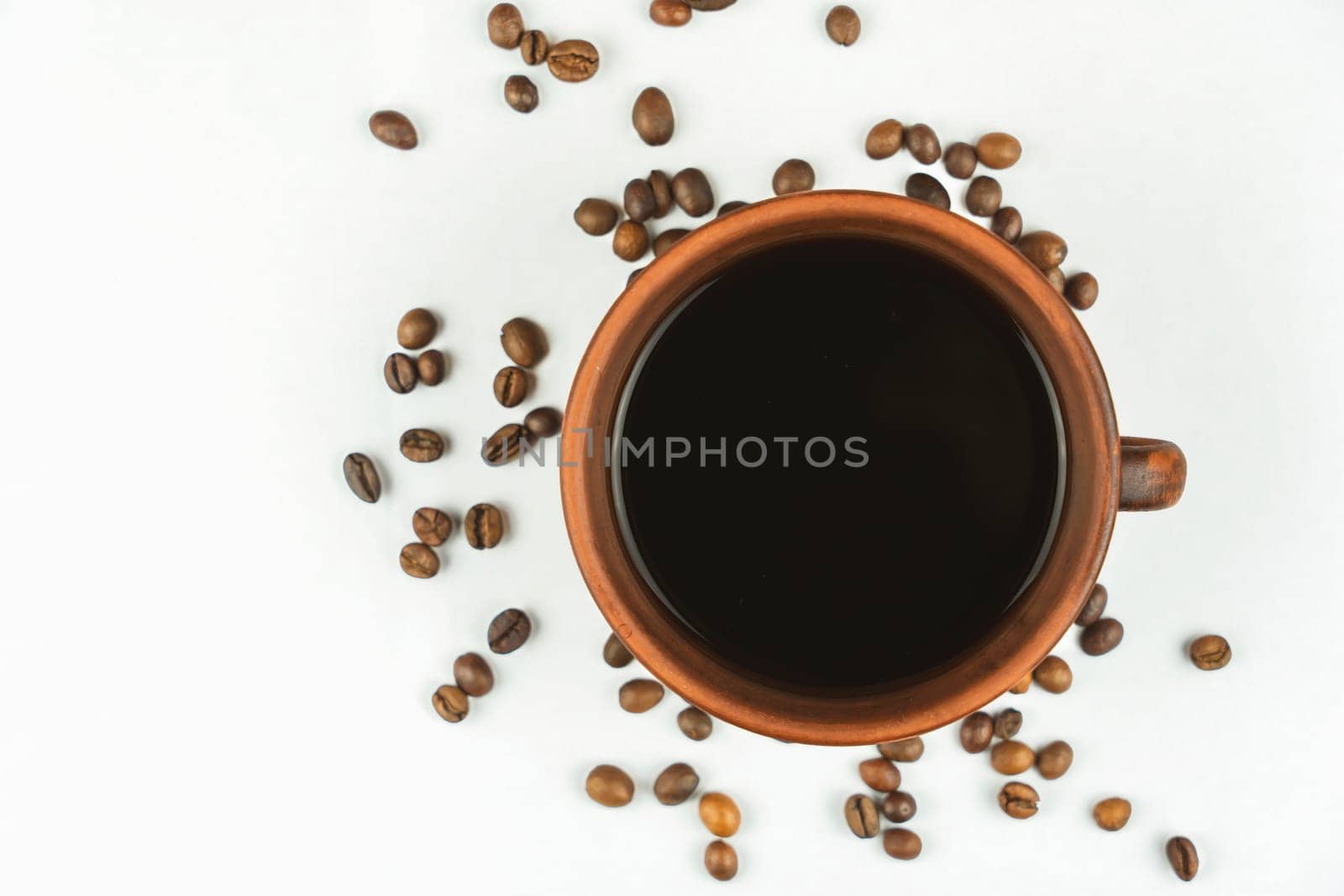 Overhead view of a ceramic coffee cup filled with black coffee surrounded by scattered roasted coffee beans on a clean white background.