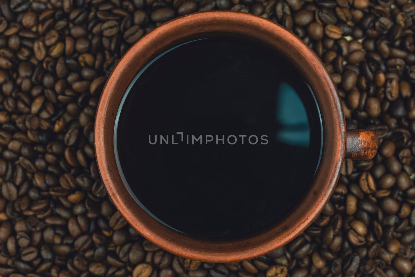 A clay cup filled with black coffee placed on a bed of roasted coffee beans, viewed from above, creating a rustic and warm composition, no people.