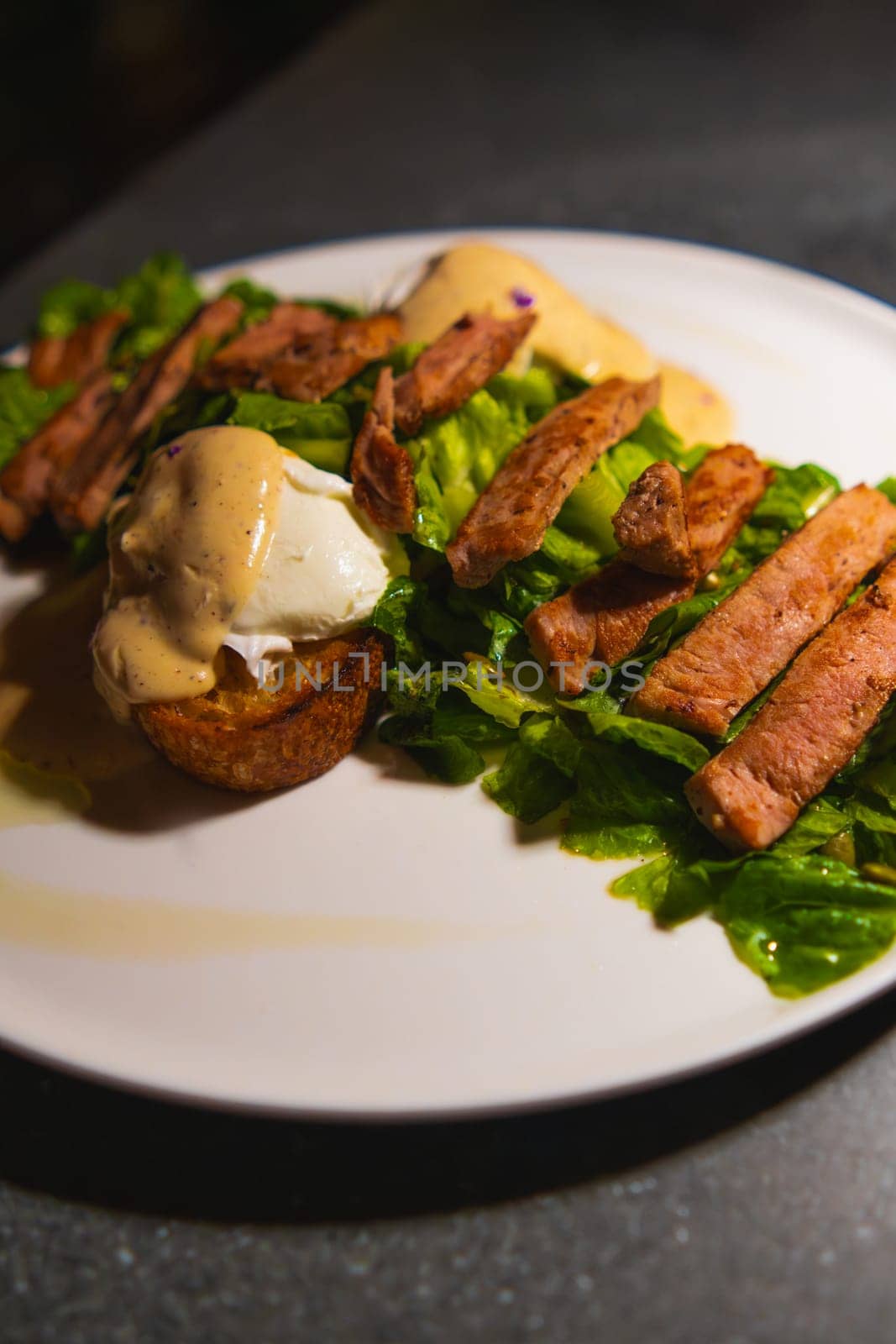 Gourmet steak salad with poached eggs and crispy bread on a white plate, dark moody lighting.