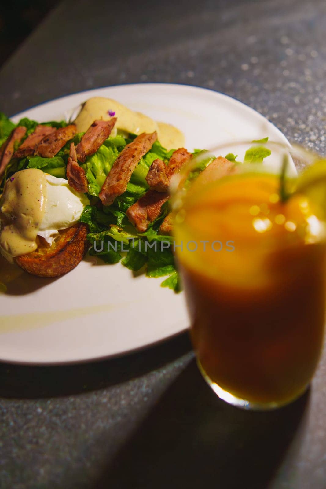 a glass of orange juice next to a plate of chicken meat and green salad on the table in the cafe at night