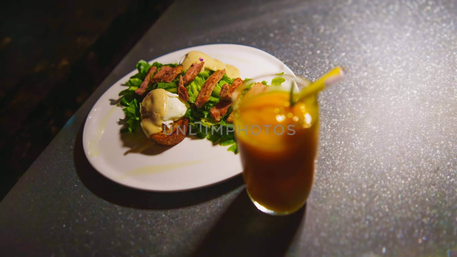a glass of orange juice next to a plate of chicken meat and green salad on the table in the cafe at night