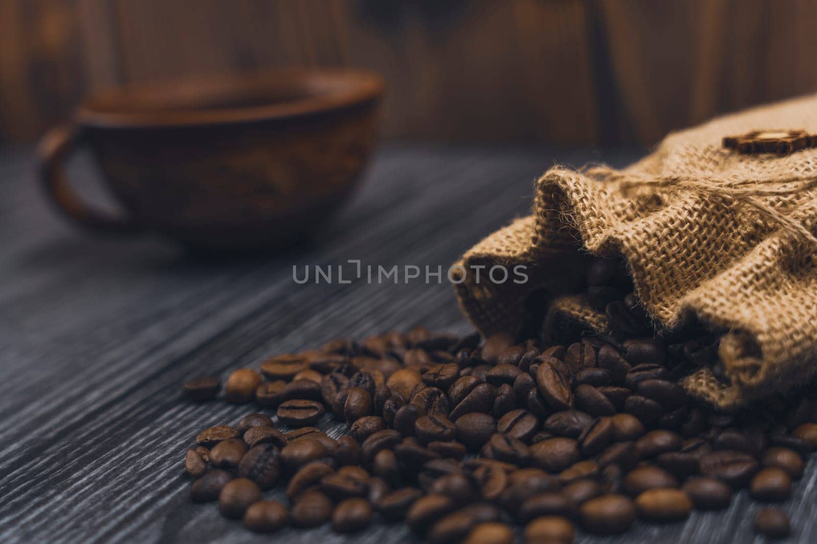Rustic burlap sack spilling roasted coffee beans onto a dark wooden surface, with a blurred ceramic coffee cup in the background, creating a cozy atmosphere.