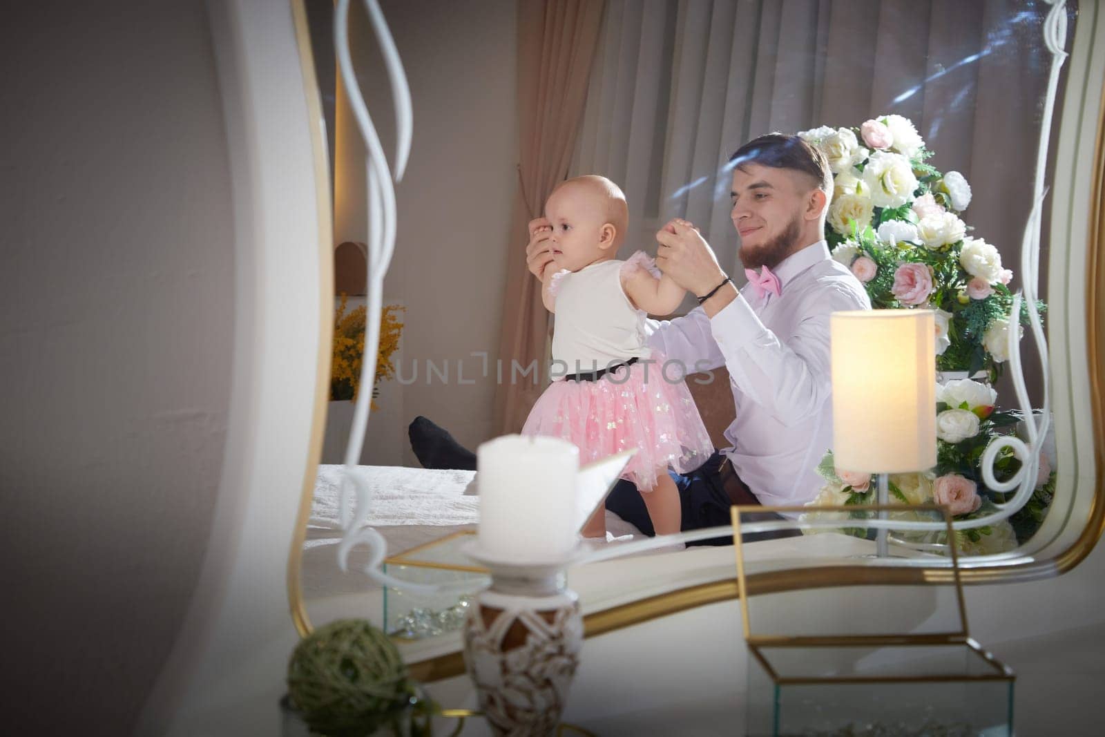 A father embraces his baby daughter near a floral arrangement in a cozy indoor setting.
