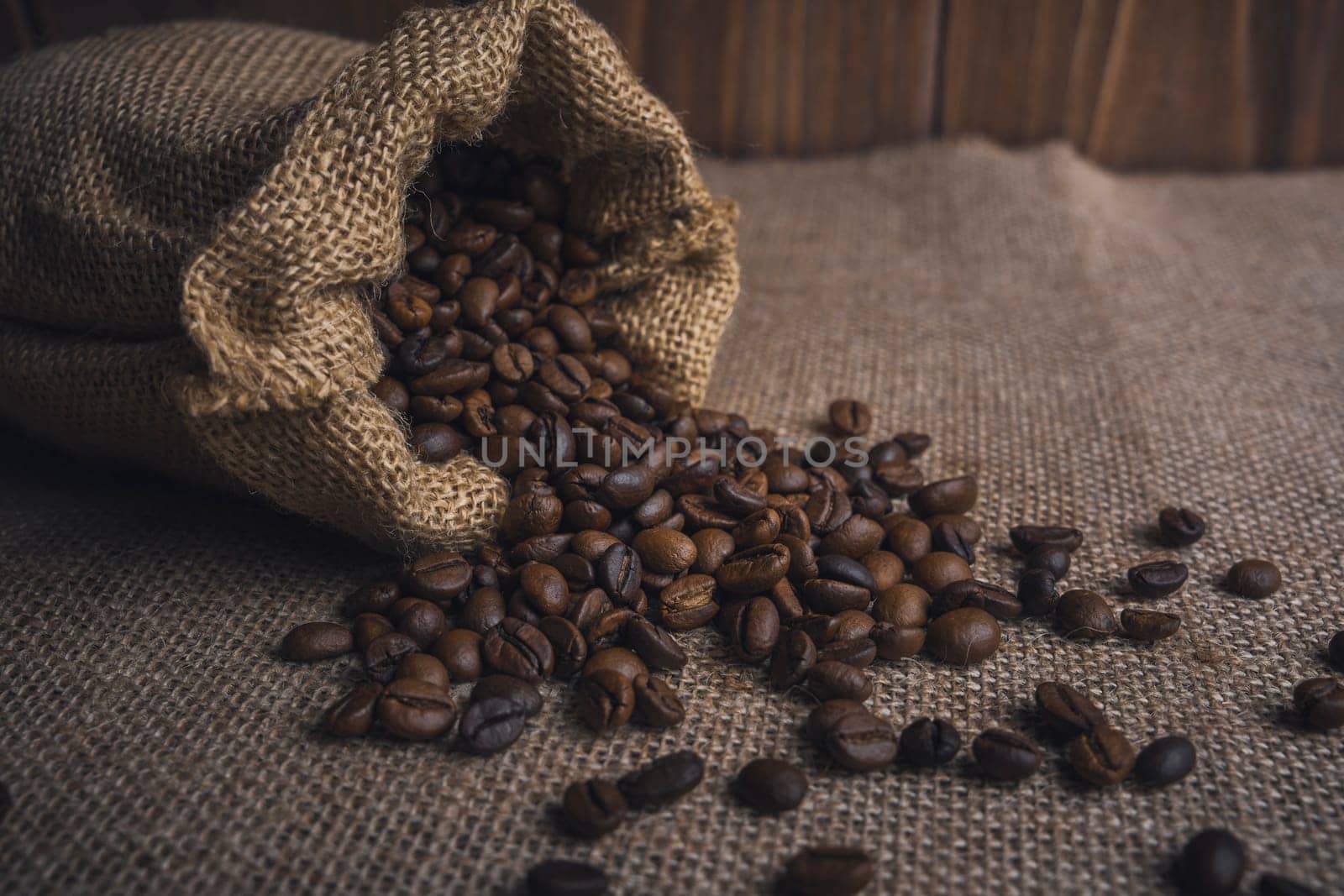 A burlap sack spilling roasted coffee beans onto a rustic burlap fabric surface with a wooden background.
