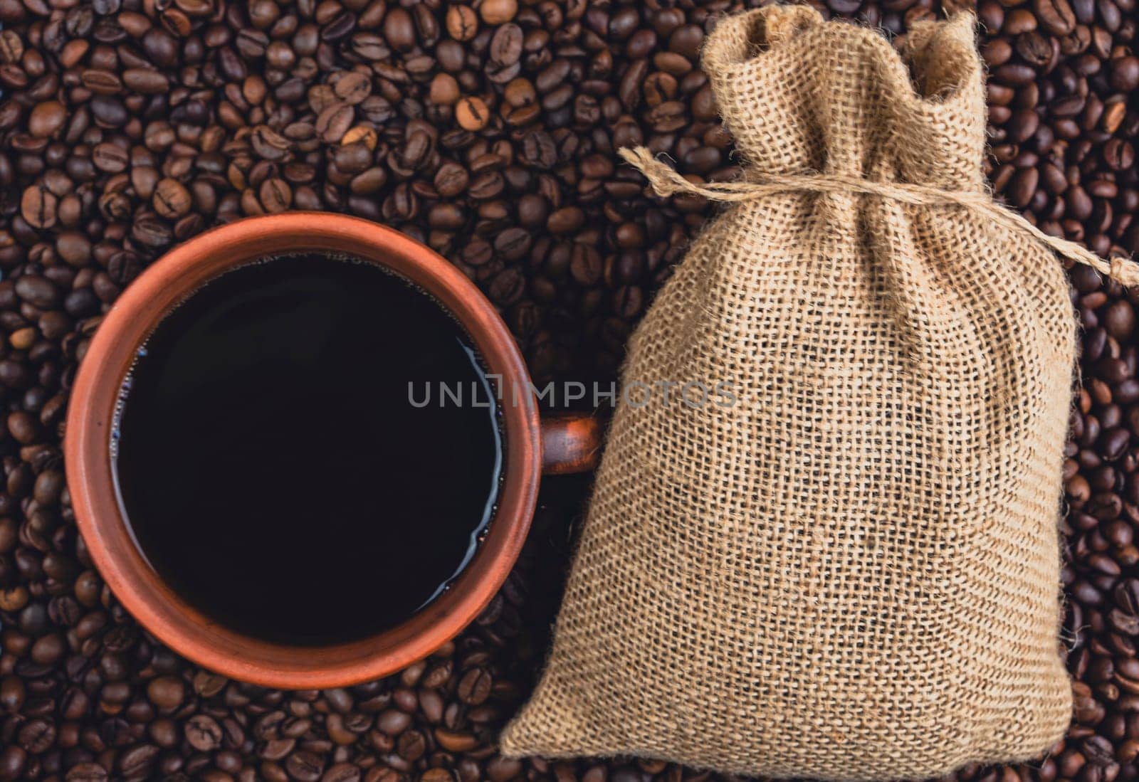 Overhead view of a cup of black coffee and a burlap sack placed on a bed of roasted coffee beans, creating a rustic coffee concept.
