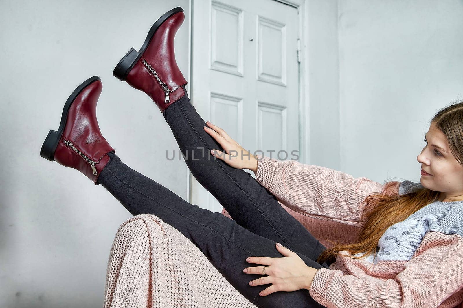 Stylish model effortlessly poses with legs extended, highlighting fashionable burgundy ankle boots in a chic room.