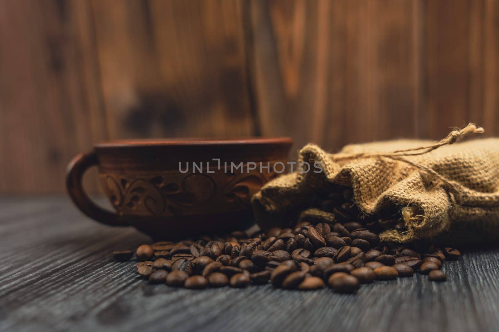 Close-up of a decorative rustic cup and a burlap sack spilling roasted coffee beans on a wooden surface, with a warm wooden background.