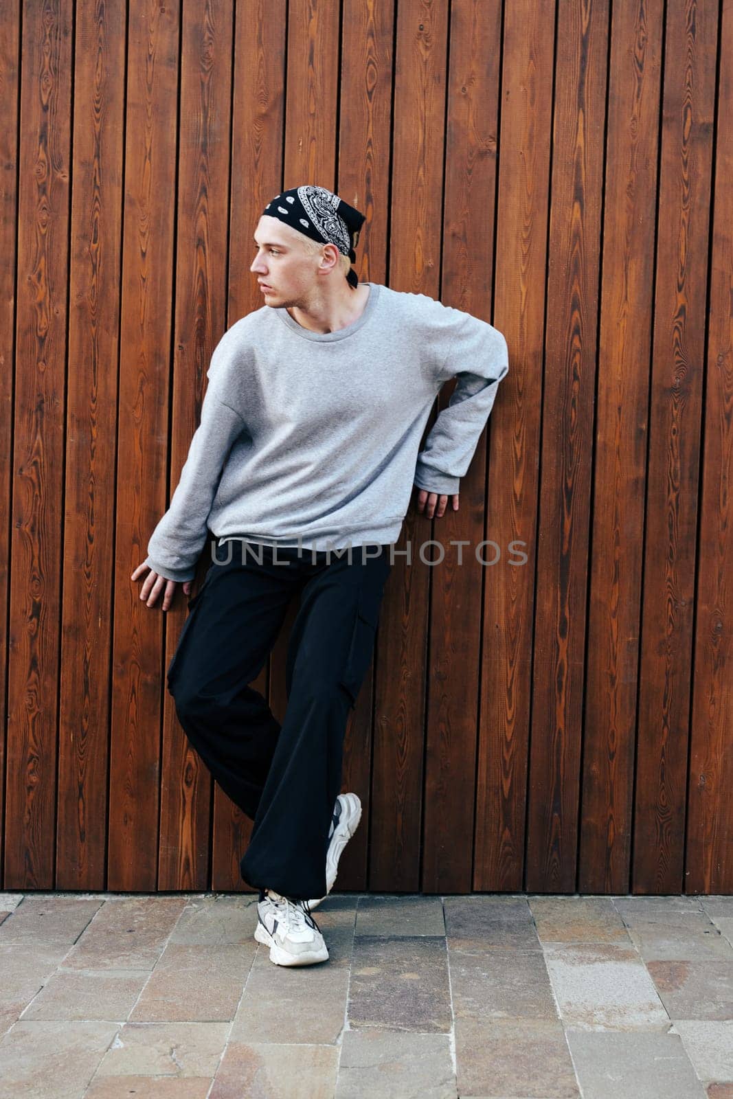 A young man in a grey sweatshirt and black pants with a black bandana on his head is leaning against a brown wooden wall.