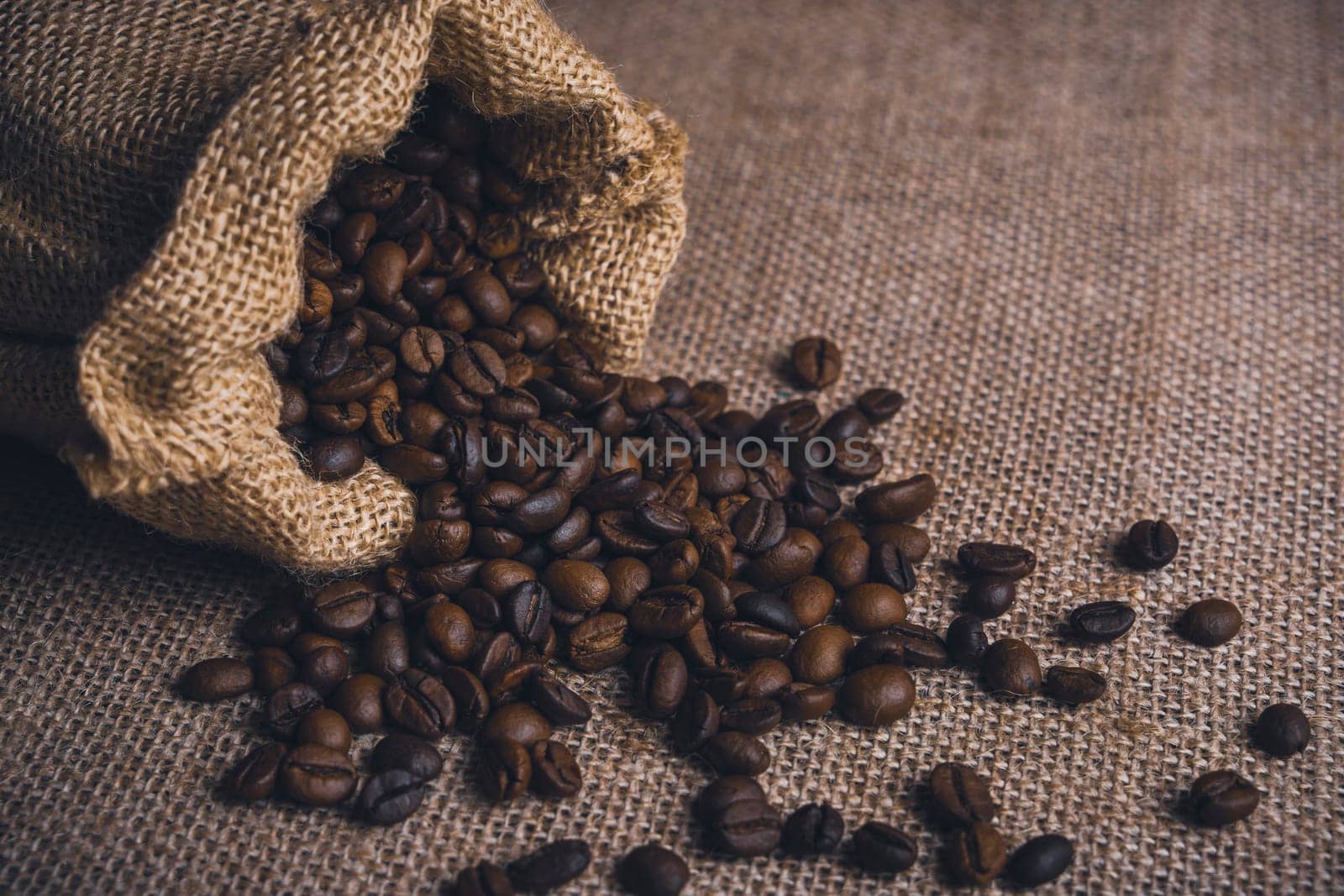 Burlap sack tipped over with roasted coffee beans spilling onto a textured burlap fabric background.