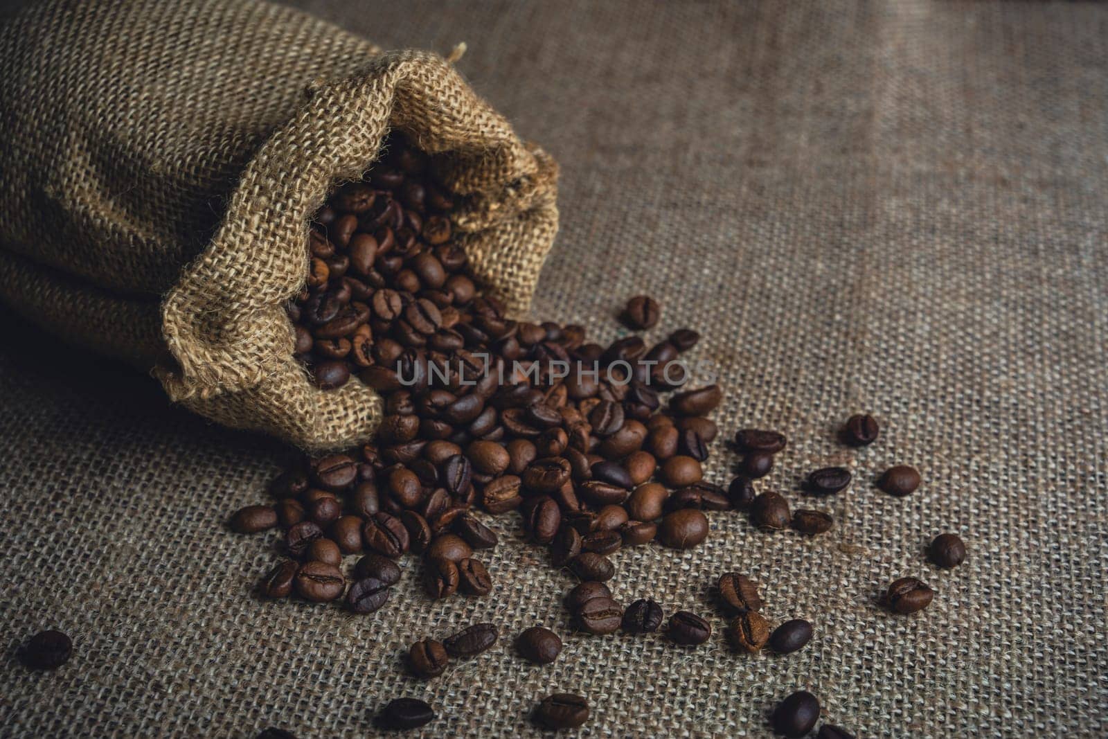 A burlap sack tipped over, spilling roasted coffee beans on a textured surface, showcasing a rustic and natural coffee theme.