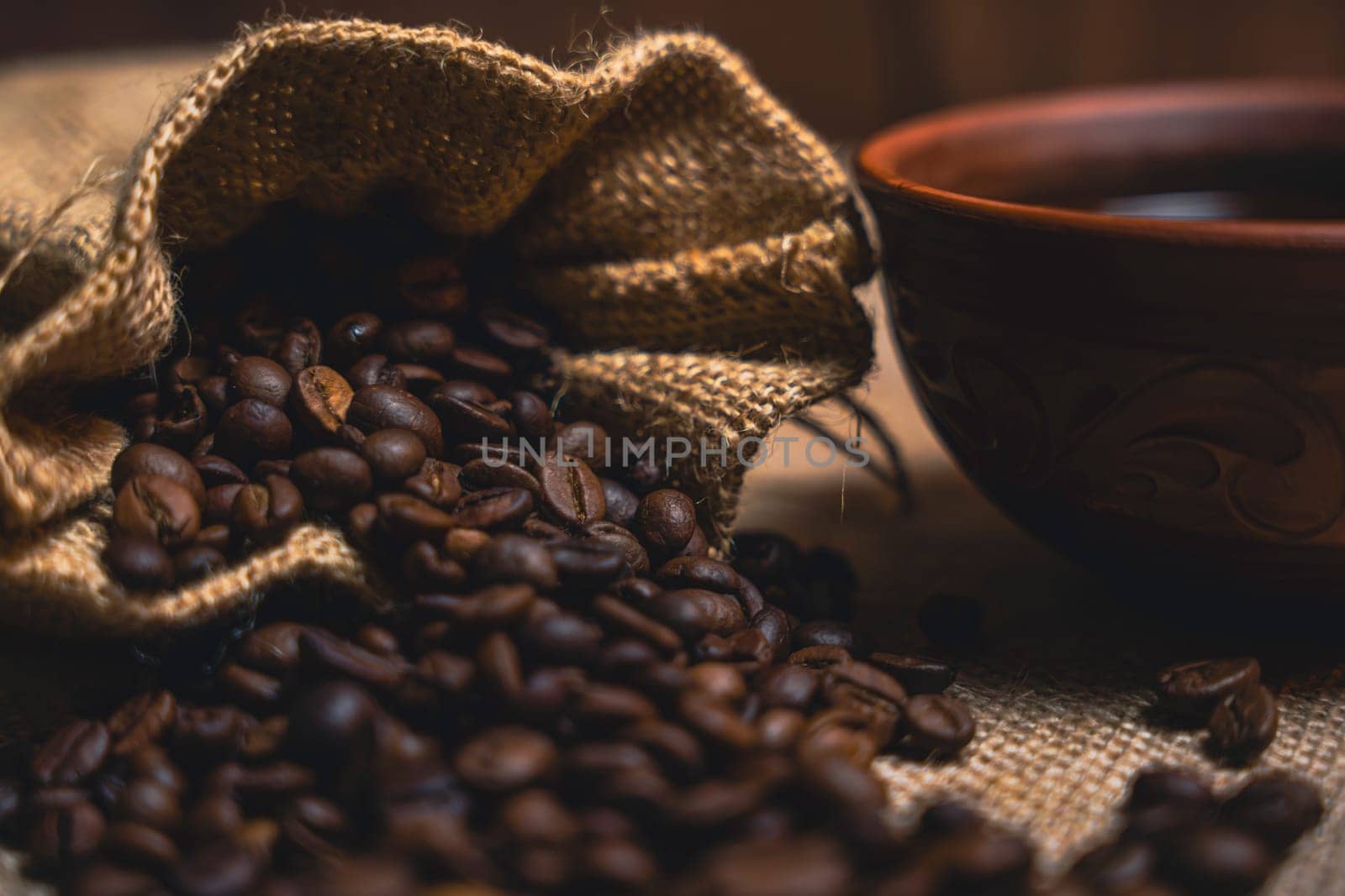 A close-up view of a burlap sack spilling roasted coffee beans next to a brown ceramic cup of black coffee on a textured burlap surface.