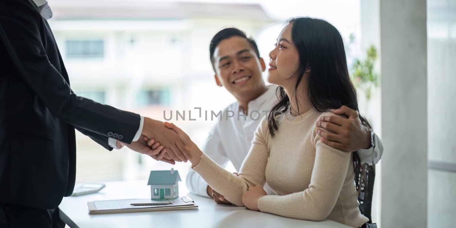 Young Asian couple making contract with house sale agency. man and his wife sitting signing the contract next to him looking the contract document with smile. real estate agreement successful concept.