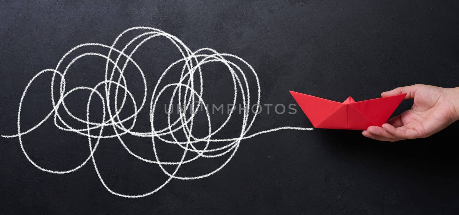 A female hand holds a red paper boat against a black chalkboard background with a long path. Concept of mentorship and assistance