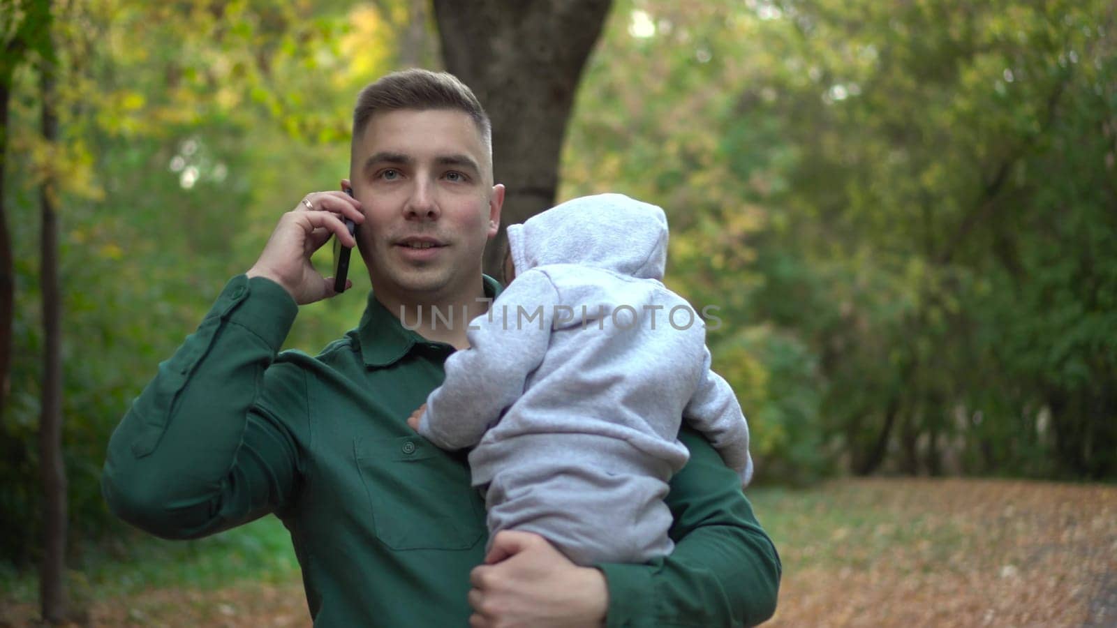 A young father holds his son in his arms and talks on the phone. A man with a child in the park with a smartphone. 4k