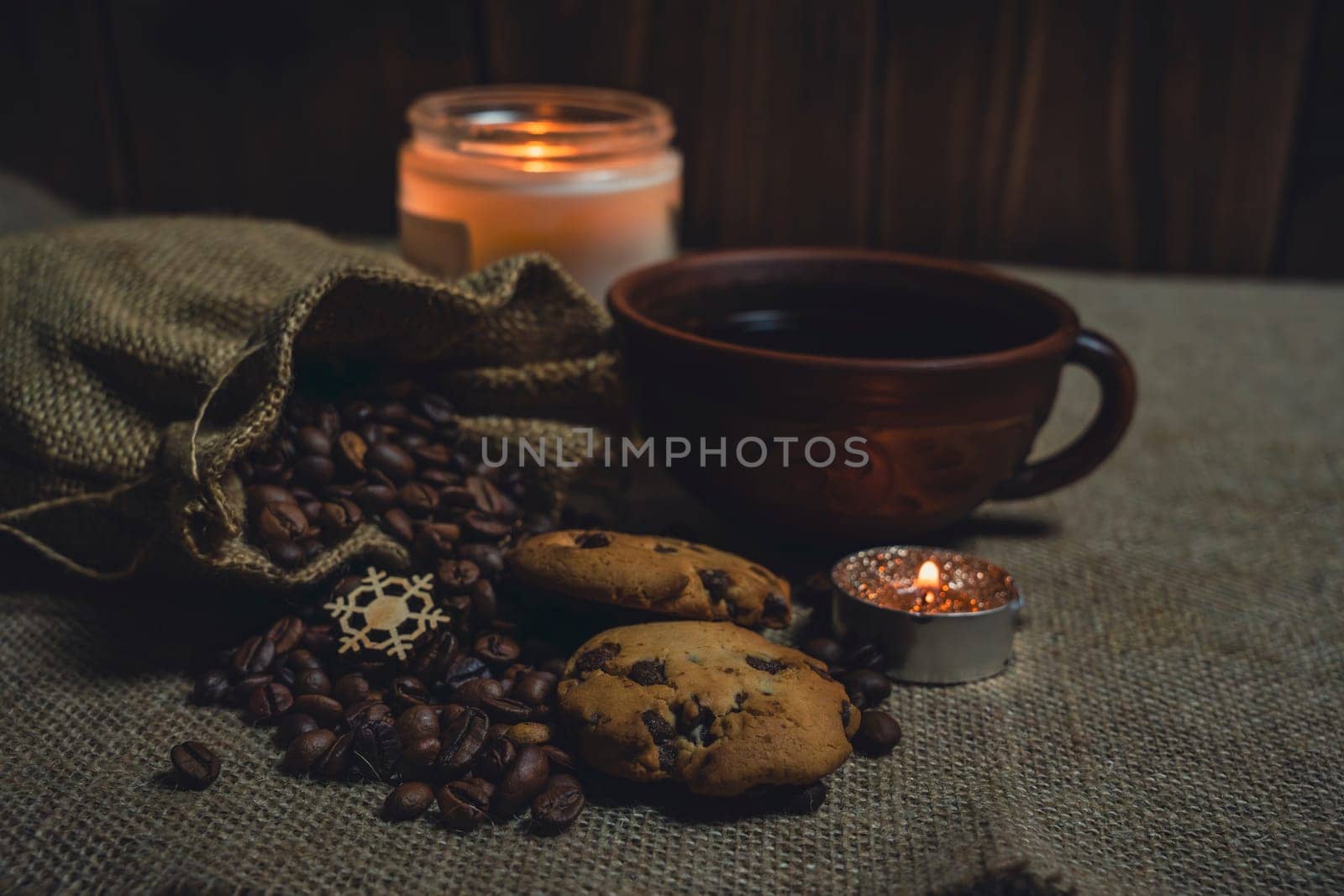 A rustic brown ceramic cup of black coffee paired with chocolate chip cookies, burlap sack spilling coffee beans, and lit candles on a burlap fabric background.