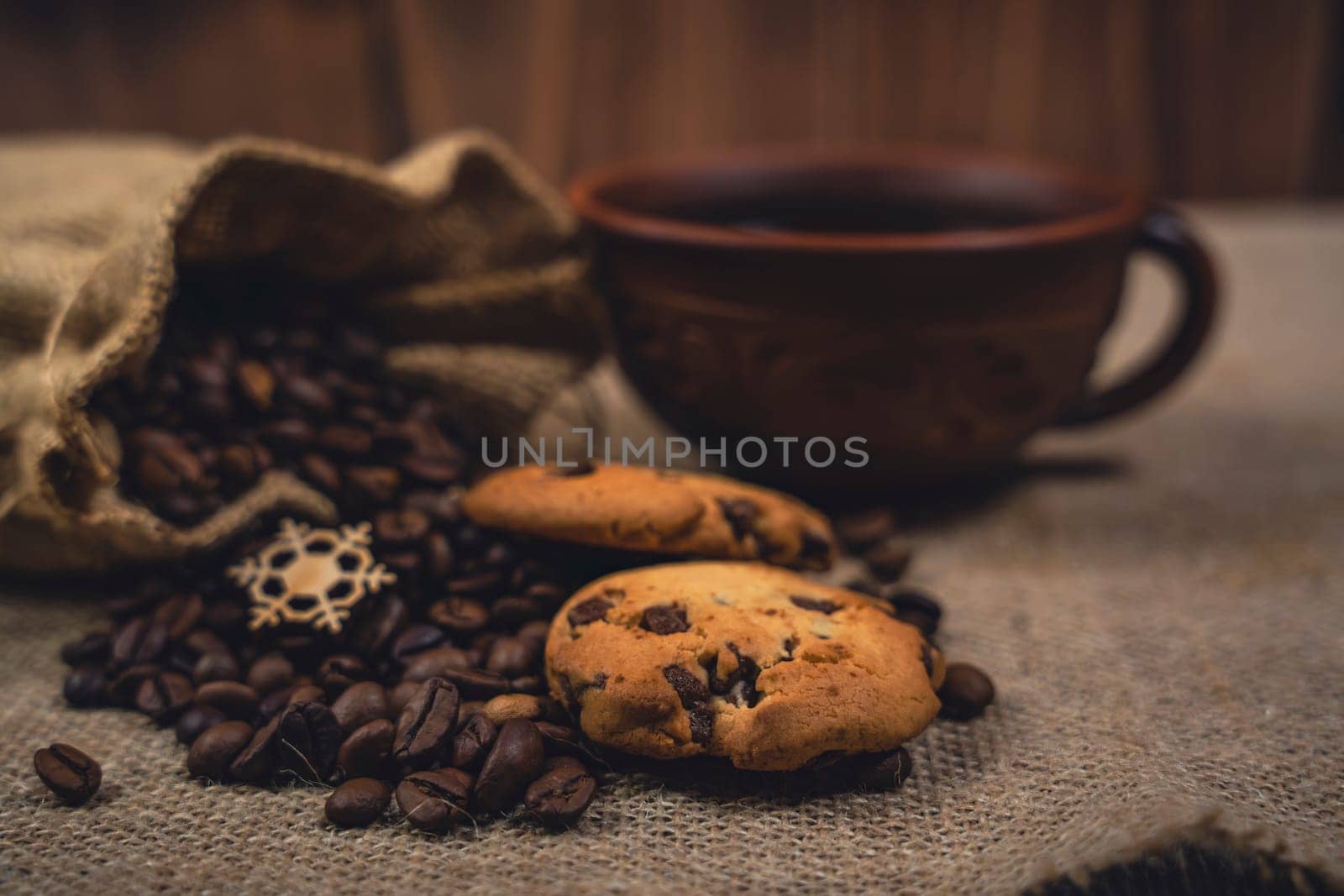 Close-up of chocolate chip cookies on burlap fabric with coffee beans spilling from a burlap sack and a rustic brown ceramic cup of black coffee in the background.