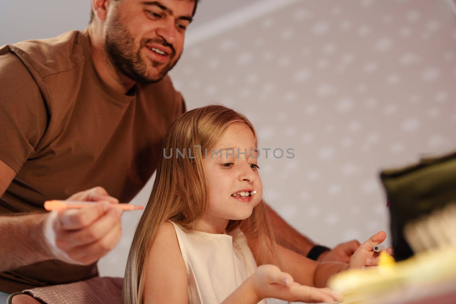A man with a beard is sitting next to a young girl. He is holding a crayon in his hand and appears to be drawing on a piece of paper.