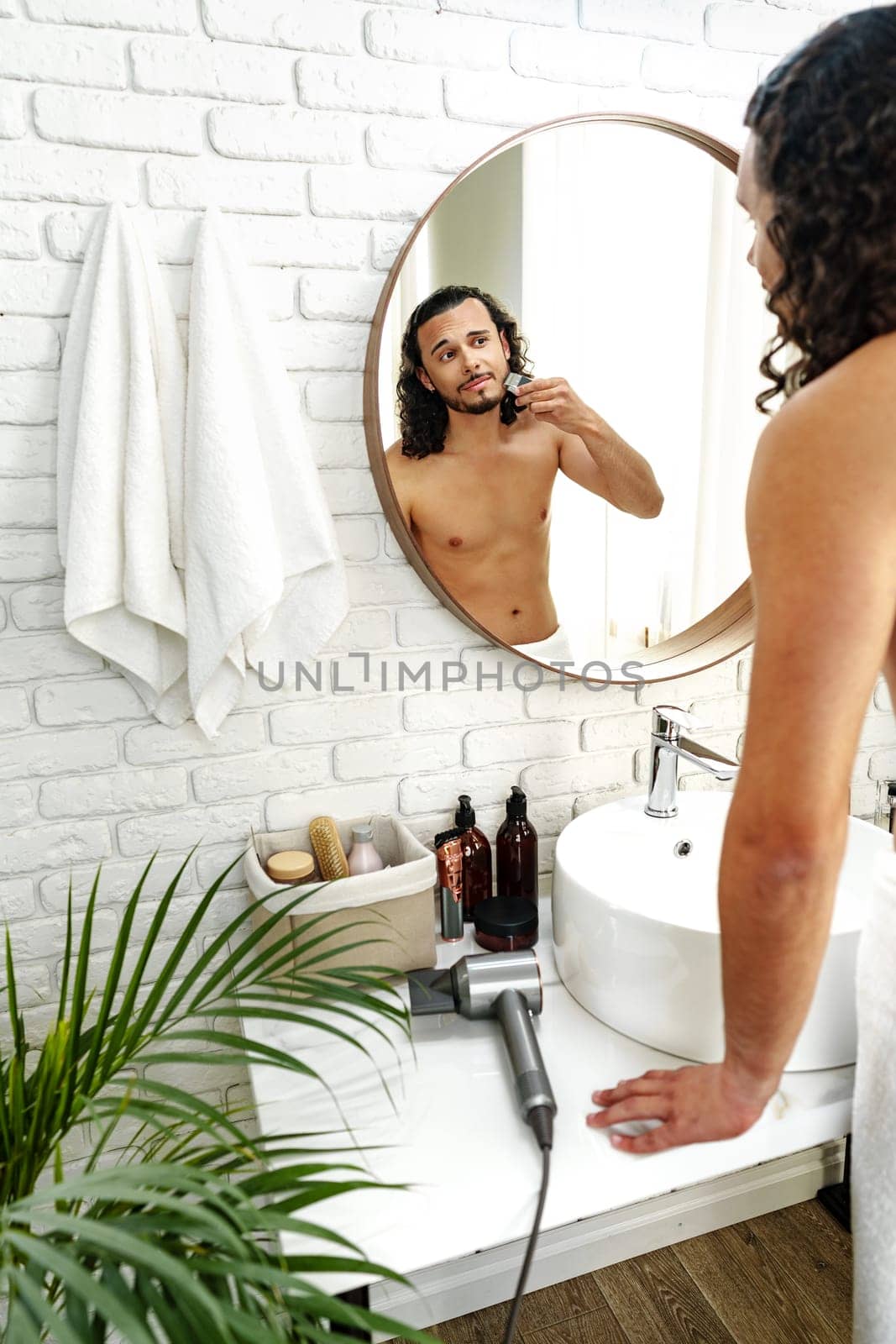 A man with curly hair grooming himself in the bathroom while looking into a round mirror close up