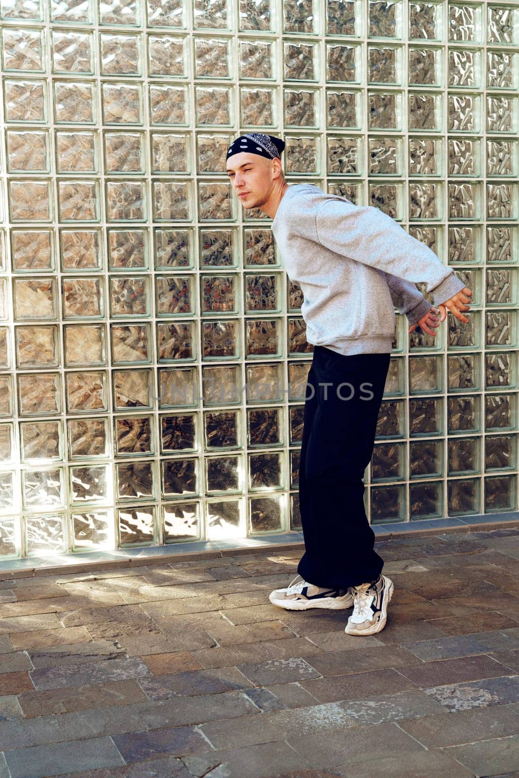 A young man, wearing a gray sweatshirt, black pants, and a bandana, stands in front of a wall made of glass blocks. The man is facing to the right, and his right hand is placed on his lower back.
