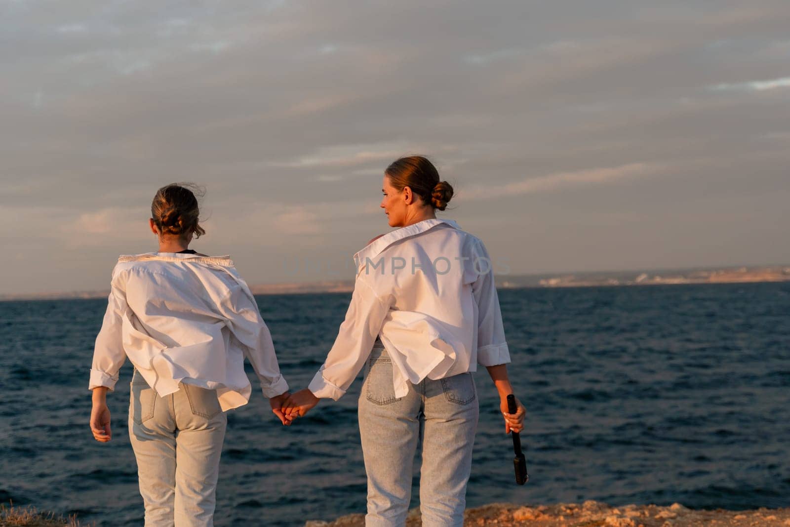 Two women, dressed in white shirts and blue jeans, walk hand-in-hand along a rocky shore, gazing at the ocean at sunset.