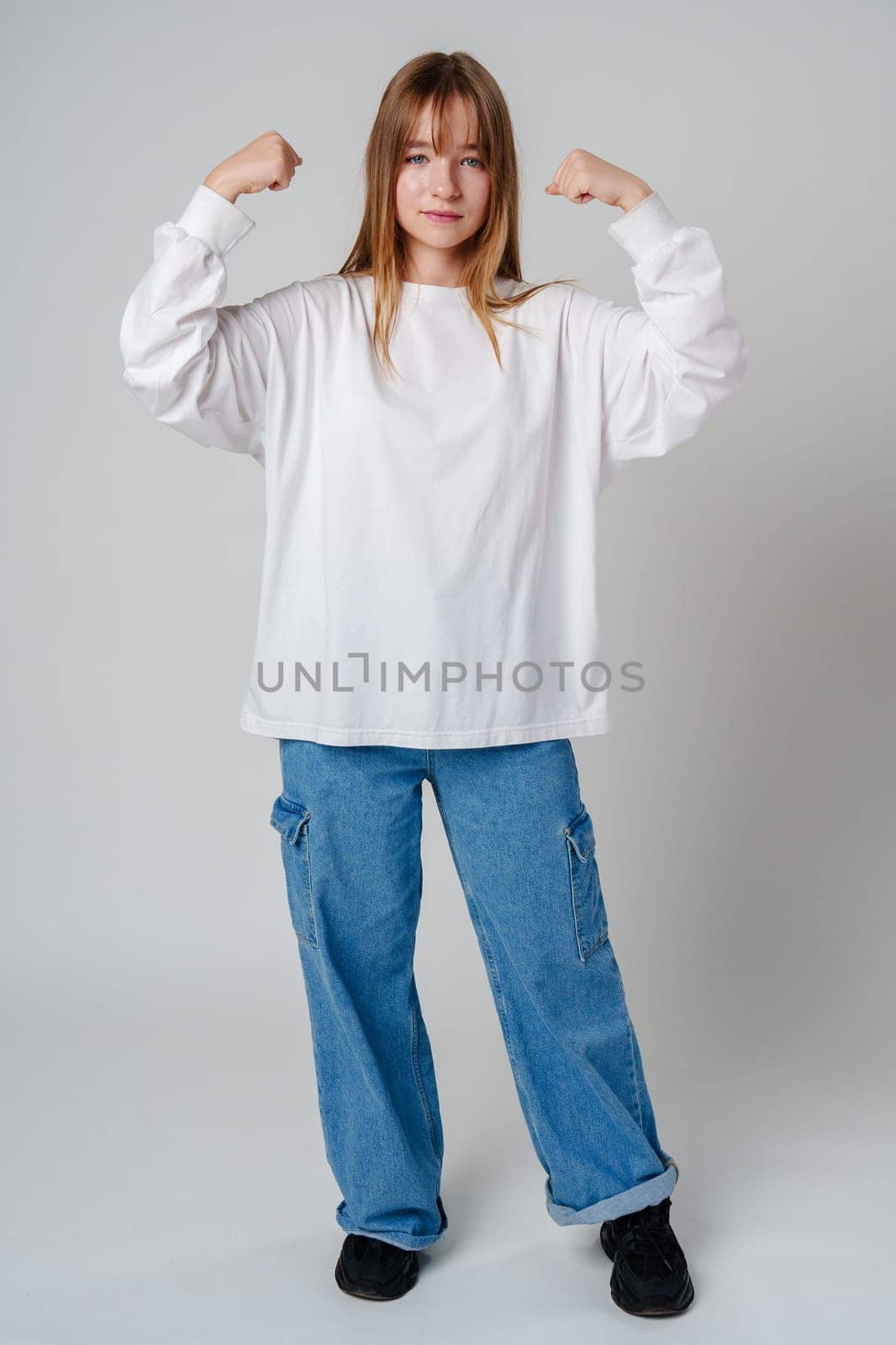 A joyful young girl with long hair wearing a white sweater stands against a plain background close up