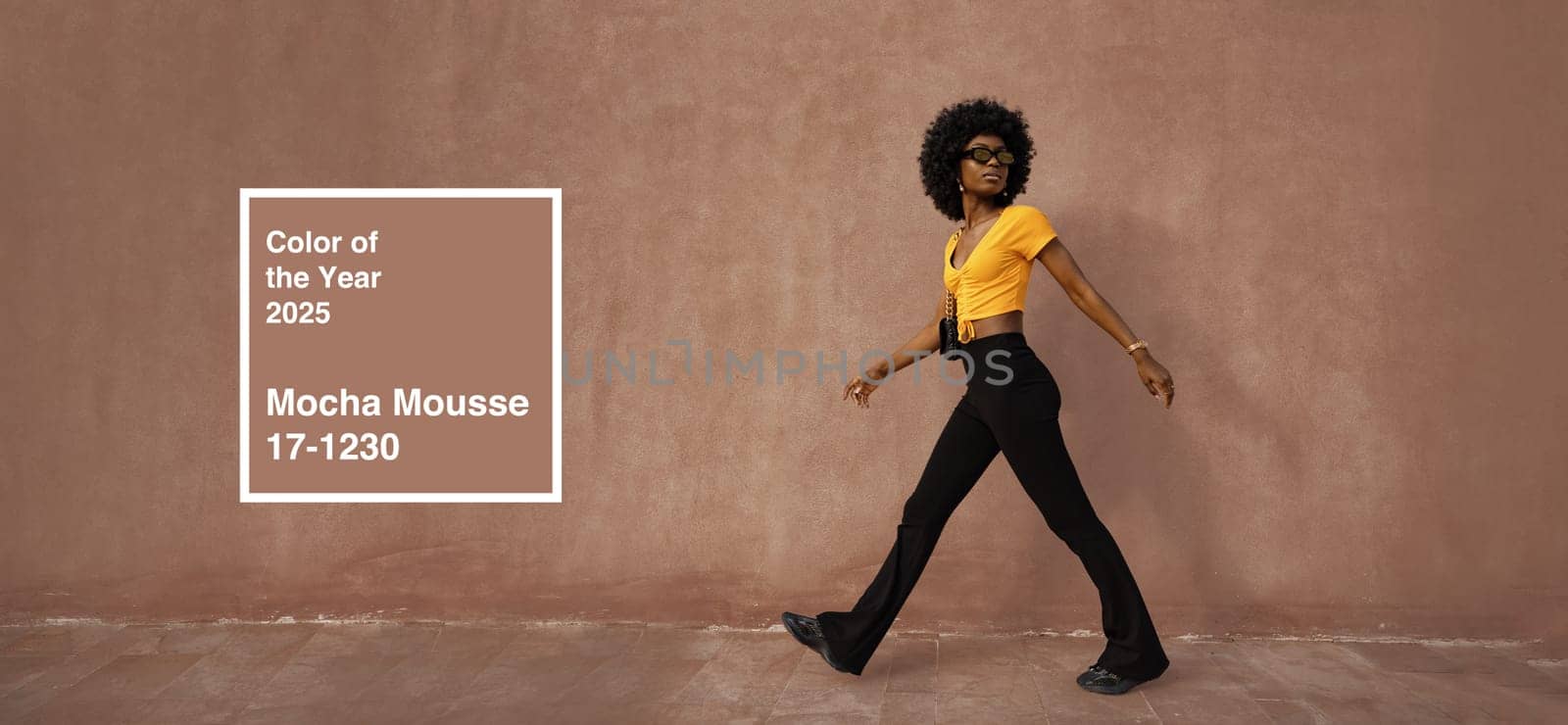 A model confidently walks against a textured backdrop wearing a vibrant yellow top and black pants. The warm light highlights the mocha mousse color trend for 2025, creating a fashionable vibe.