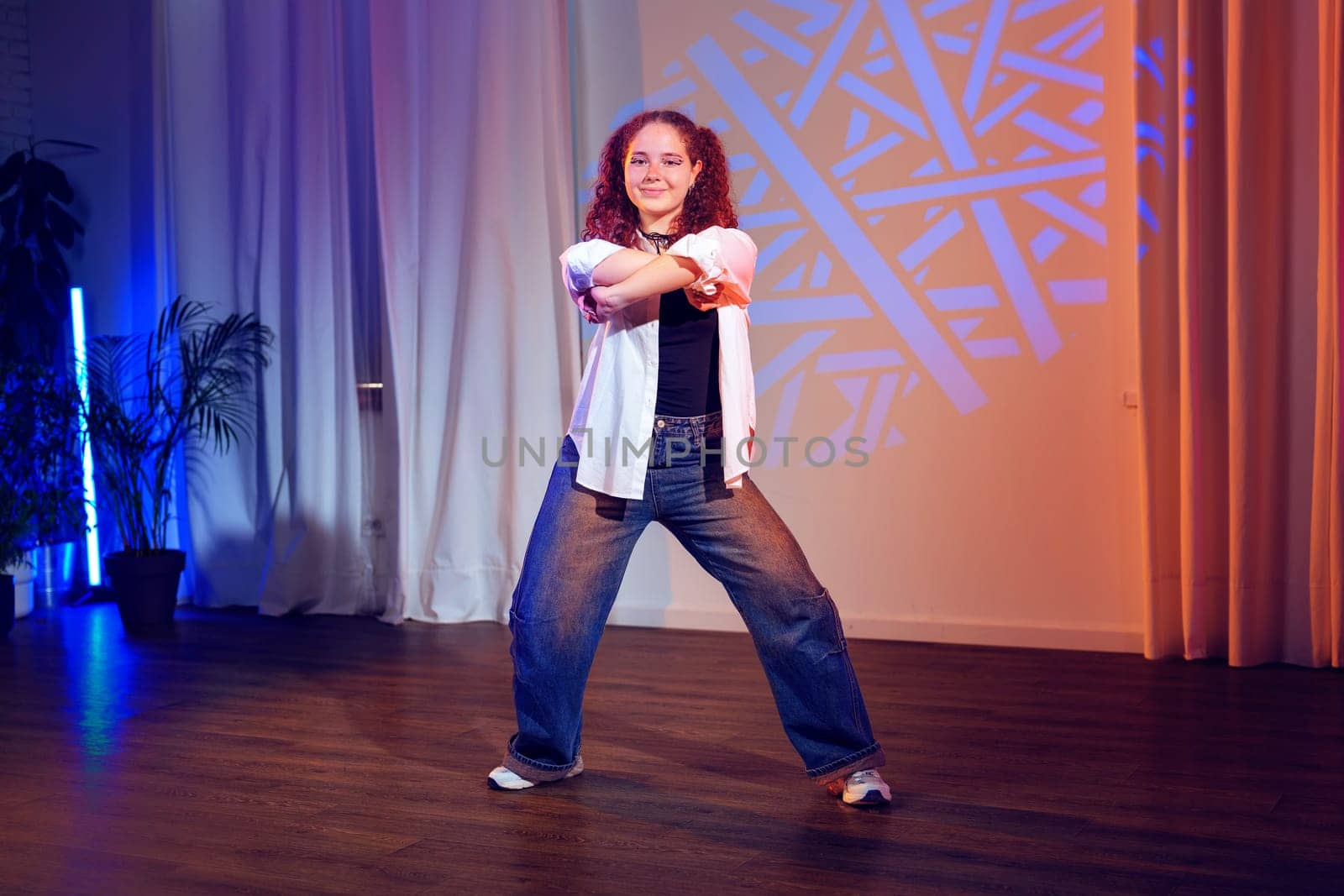 A young woman with curly brown hair is dancing in a studio. She is wearing a white shirt, blue jeans, and white sneakers.