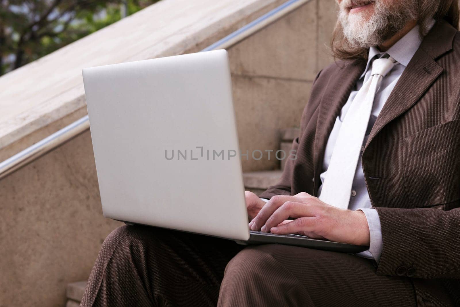 close up of an unrecognizable mature male entrepreneur in formal clothes sitting on urban stairway and working online on laptop in city, concept of business and remote work